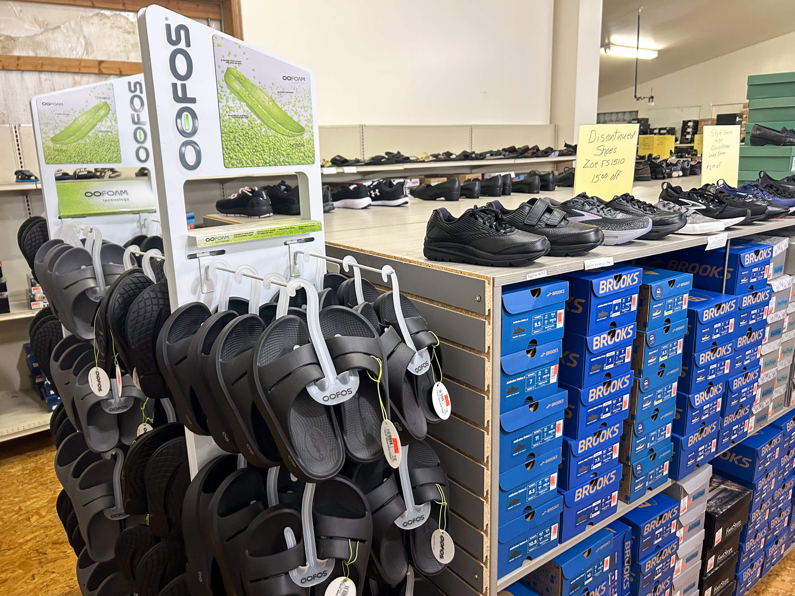 Miller's Country Store Grabill IN Display of OOFOS sandals on a rack next to shelves with various athletic shoes and stacked blue Brooks shoe boxes in a shoe store.