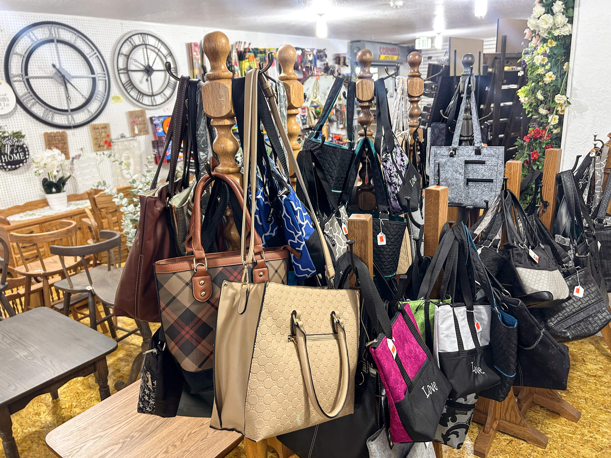 Miller's Country Store Grabill IN A display of various handbags and totes hangs from wooden posts in a store, with tables, chairs, and wall clocks visible in the background.