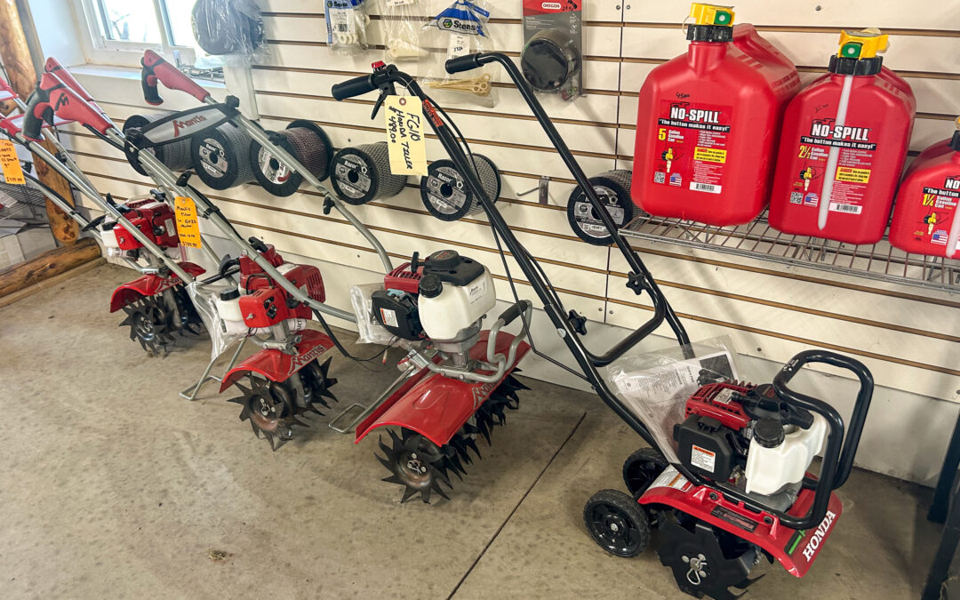 Miller's Sales & Service Centerville, MI Four gas-powered tillers are displayed on the floor in front of a wall with accessories and red fuel cans in a hardware store.