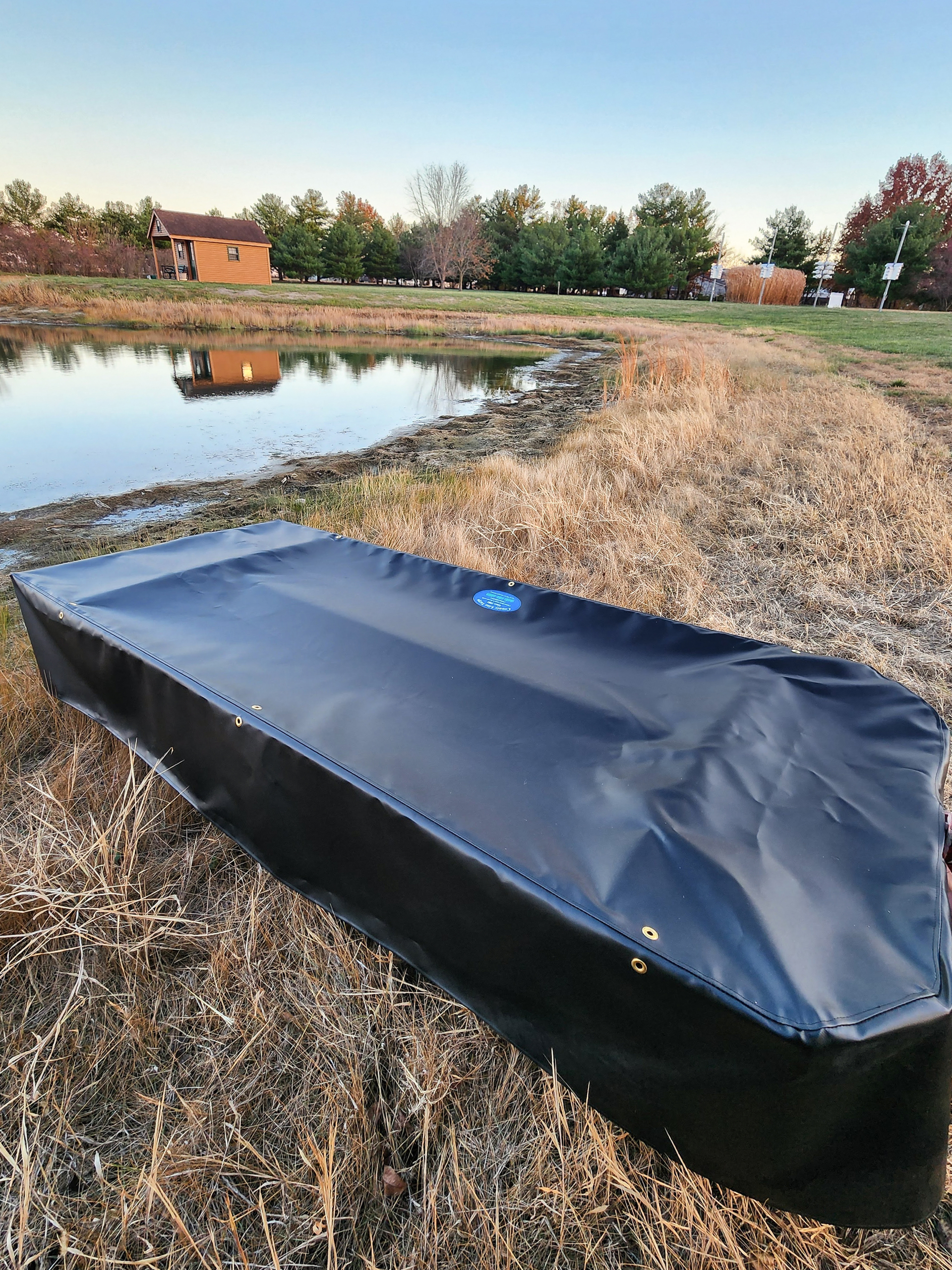 County Line Tarps Jamesport MO A black rectangular object with a fitted cover sits on dry grass near a small pond, with trees and a wooden building in the background.
