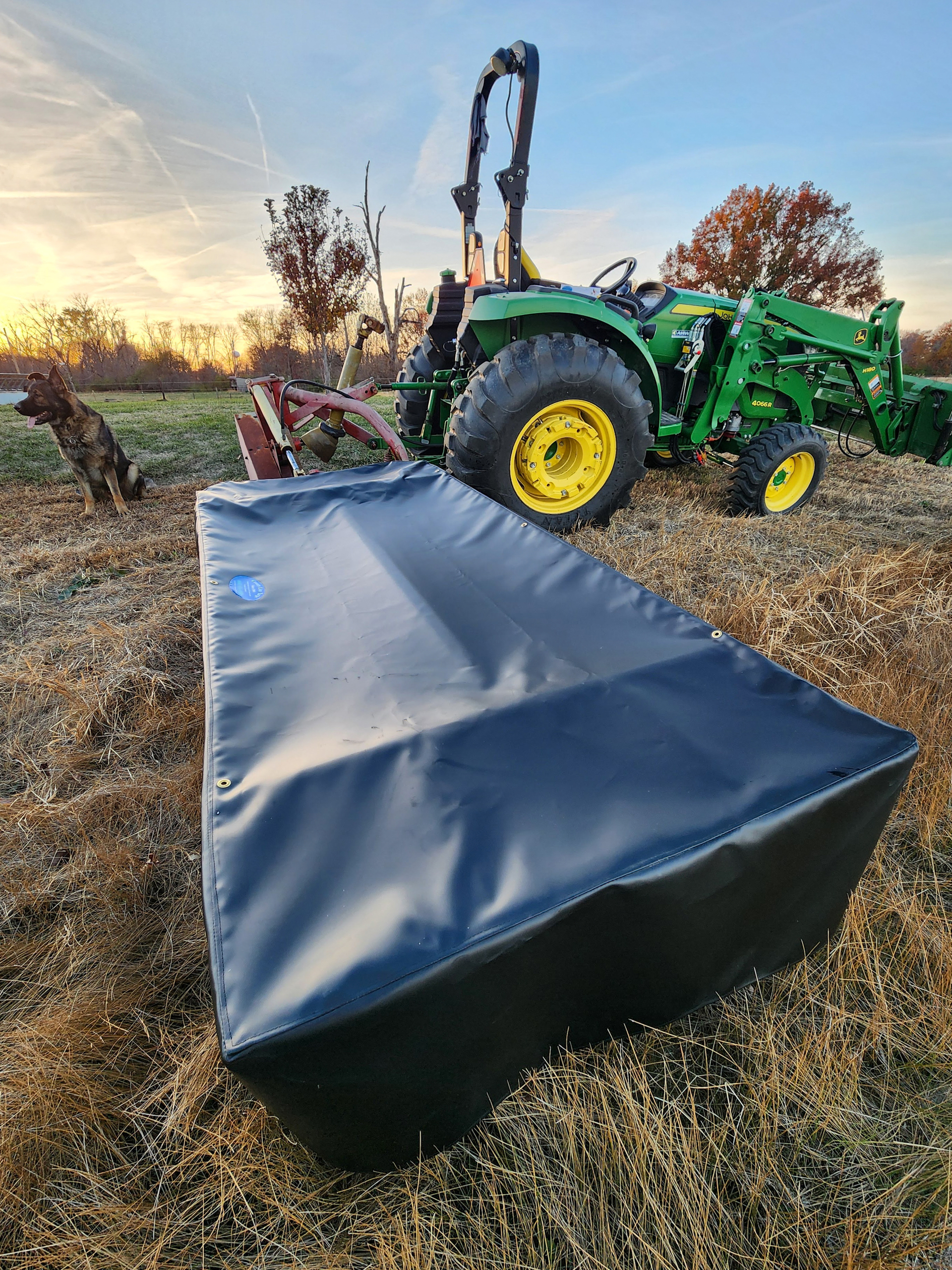 County Line Tarps Jamesport MO A green tractor with a large covered attachment is parked on dry grass. A dog sits nearby, and trees are visible in the background near sunset.