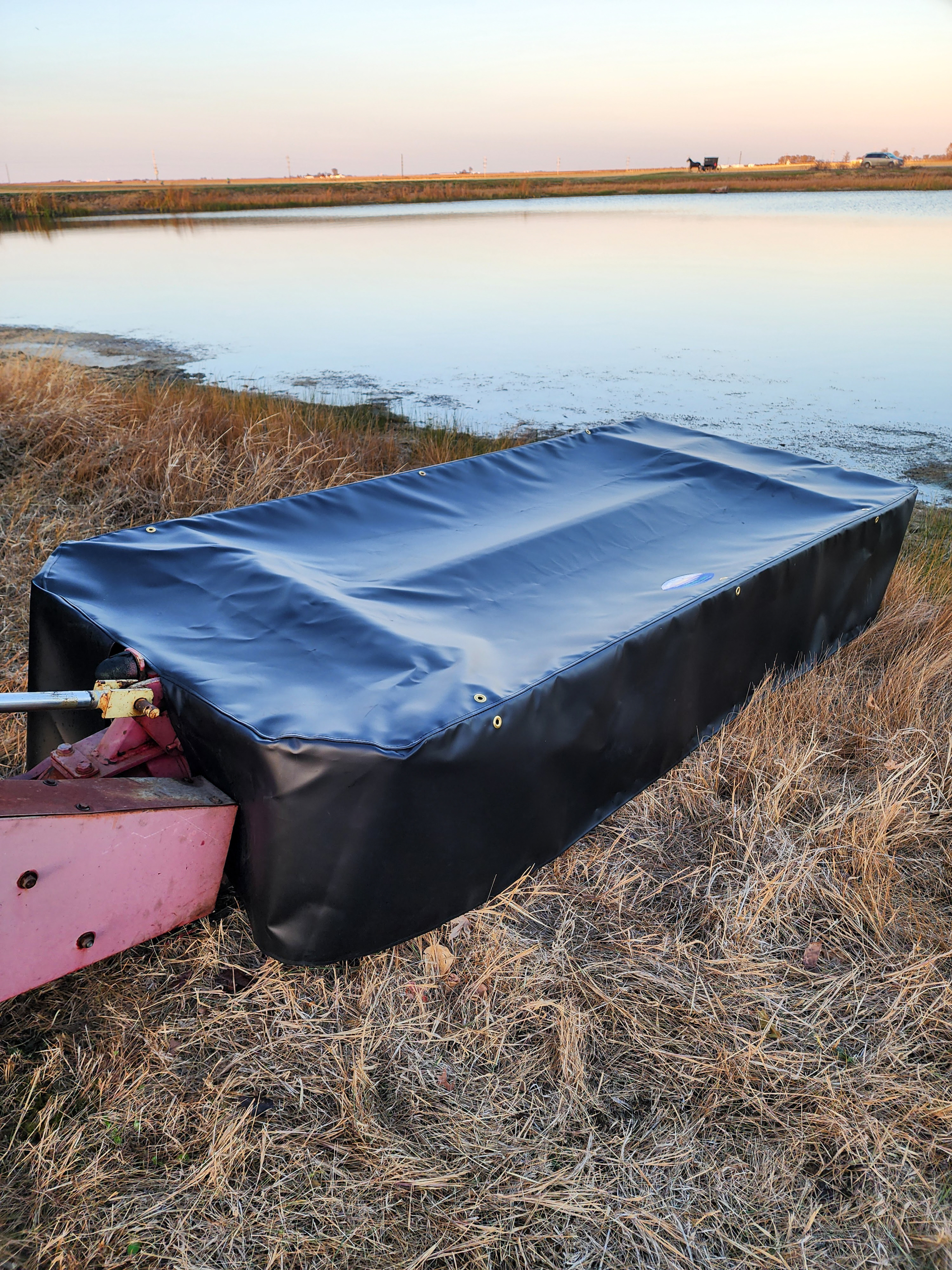 County Line Tarps Jamesport MO A black rectangular equipment cover is placed over a piece of machinery on dry grass near a pond, with open fields and a distant structure in the background.