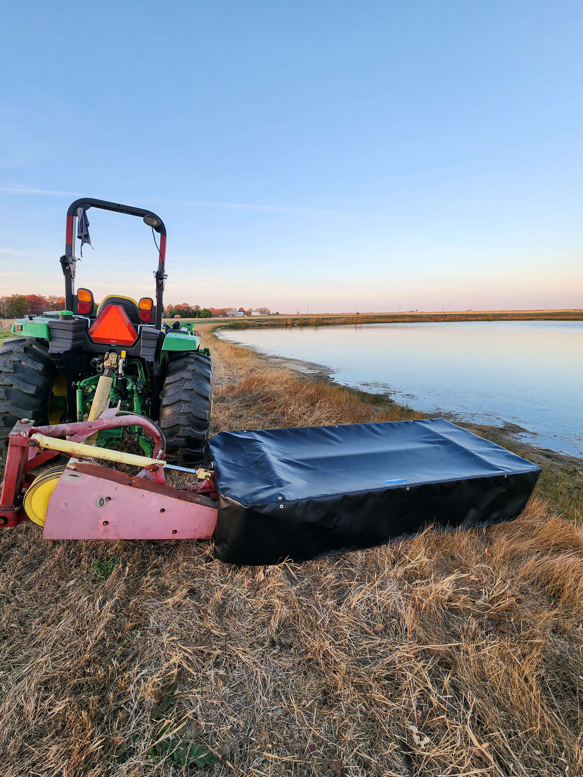 County Line Tarps Jamesport MO A green tractor with a mowing attachment stands on dry grass near the edge of a calm body of water under a clear sky.