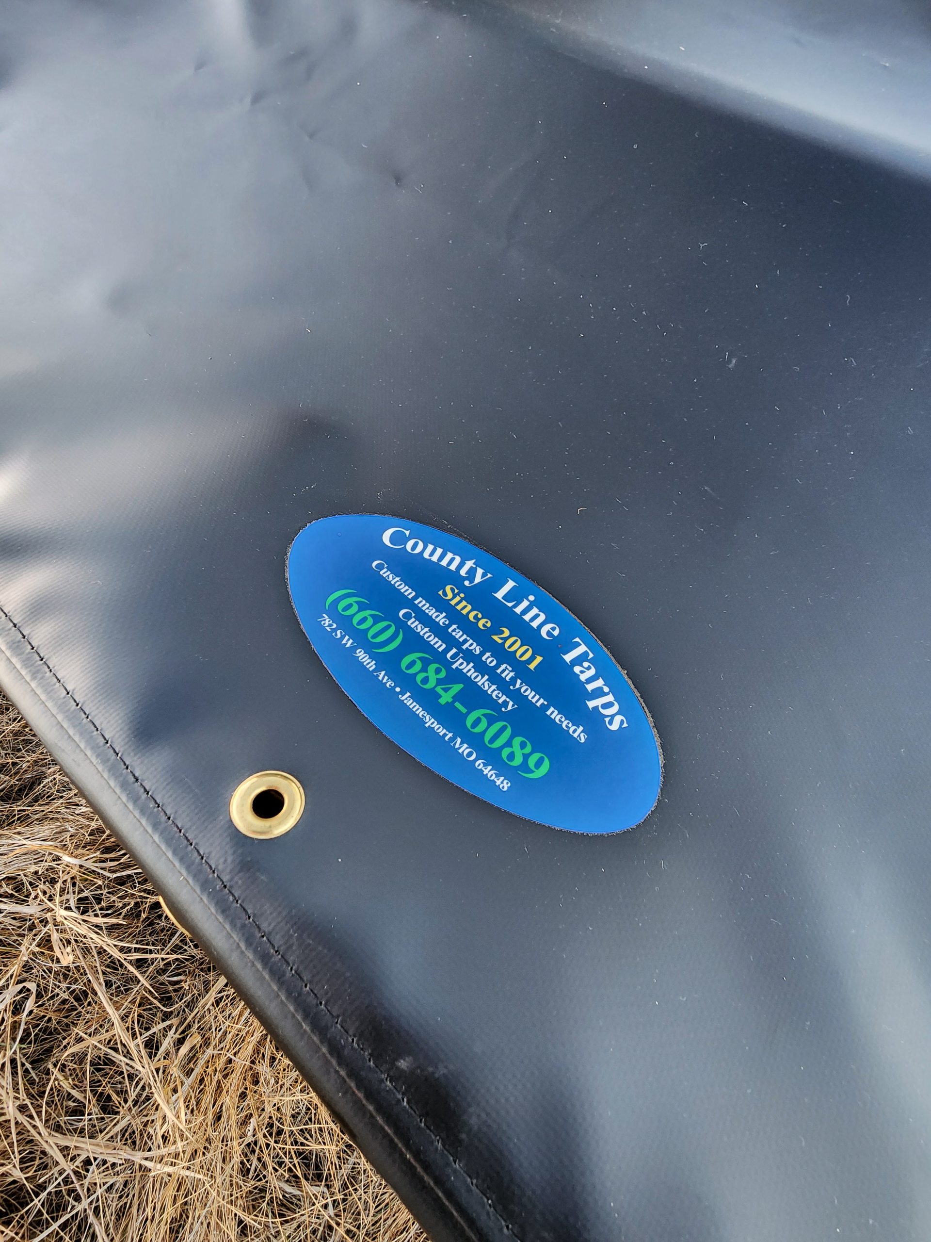 County Line Tarps Jamesport MO A close-up of a black tarp with an oval blue "County Line Traps" label and a brass grommet, lying on dry grass.