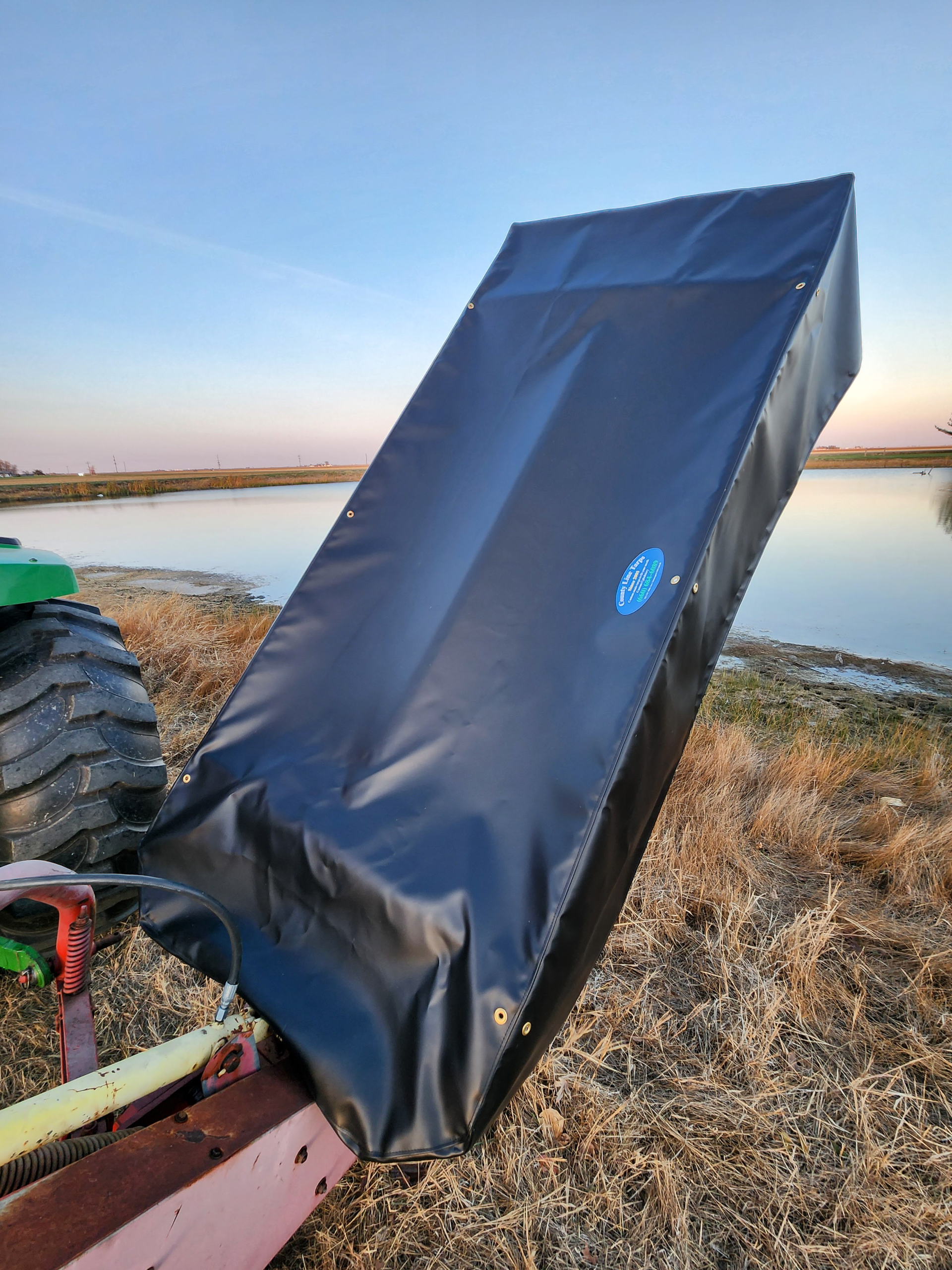 County Line Tarps Jamesport MO A black protective cover is fitted over a piece of agricultural equipment near a pond, with dry grass and a tractor tire visible beside it.