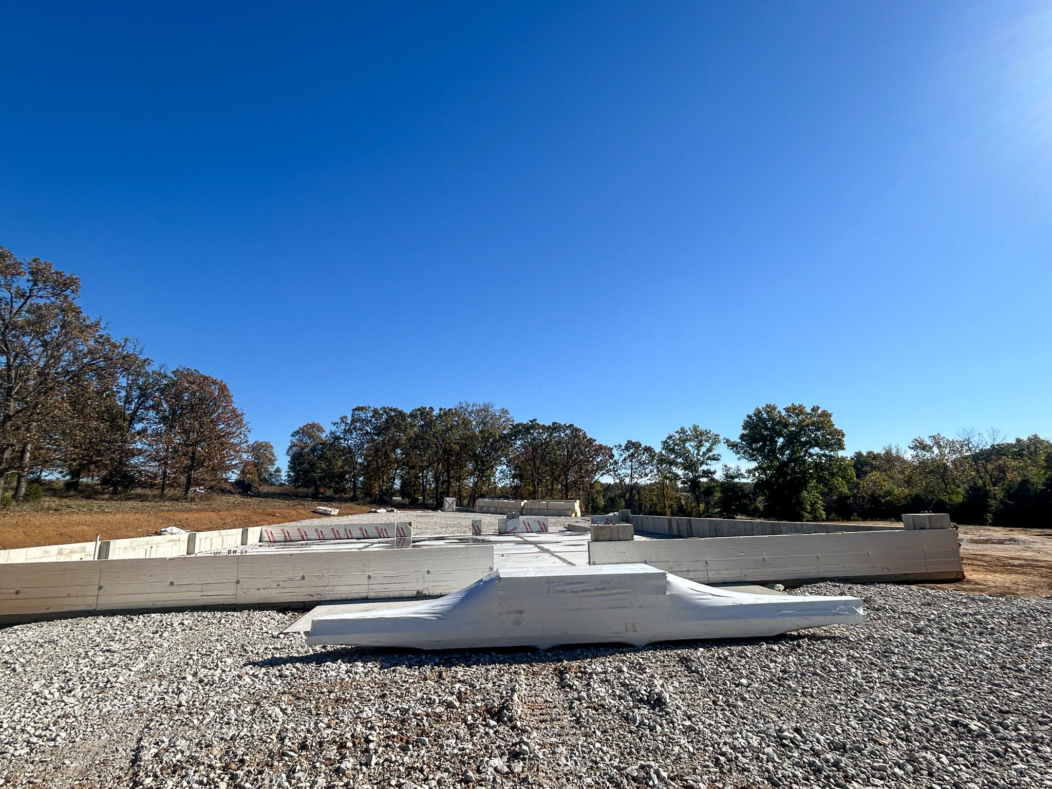 A construction site with concrete foundation forms and gravel, surrounded by trees under a clear blue sky.