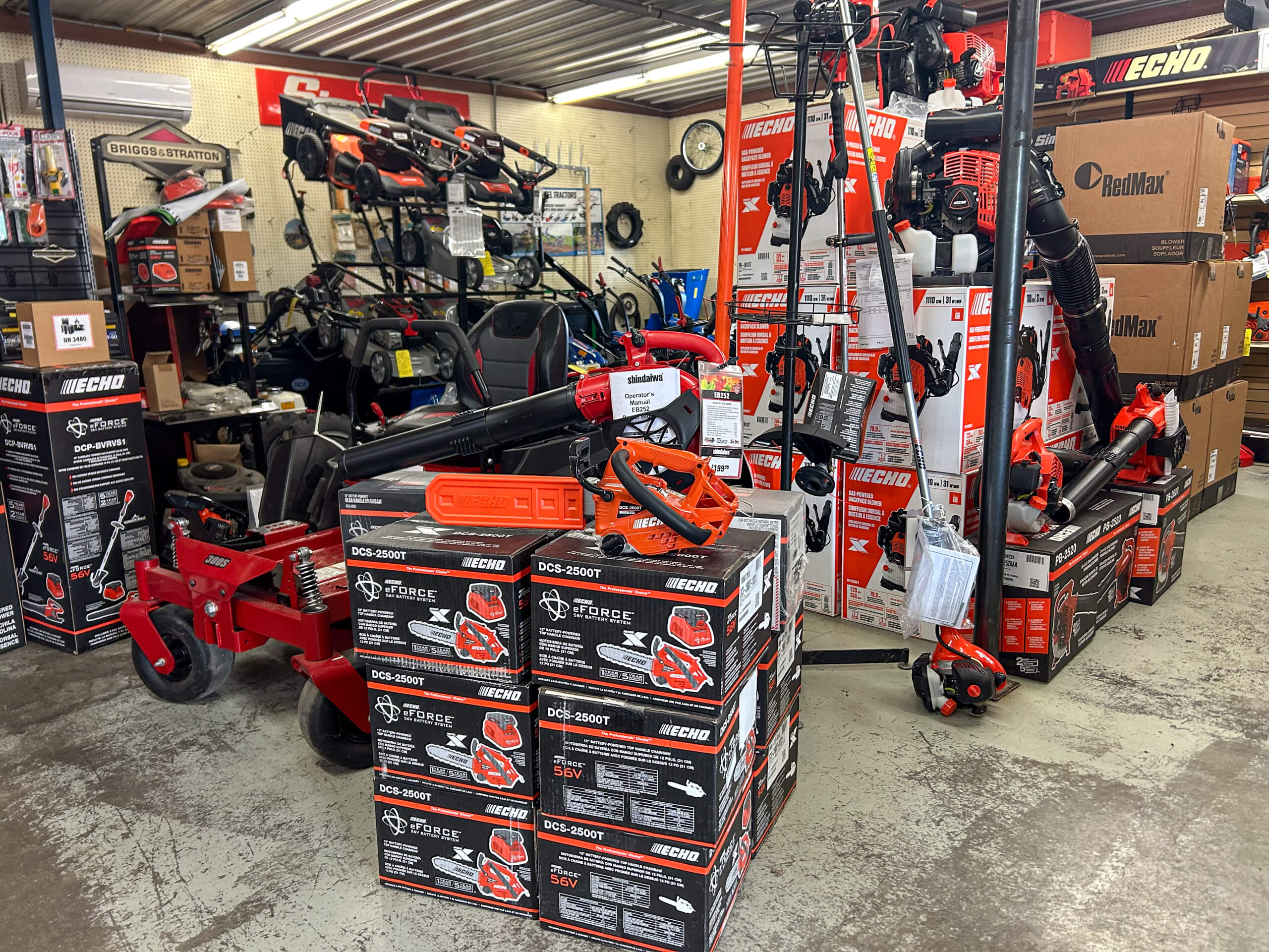 AK Small Engines Versailles, MO A hardware store displays stacked boxes of Echo outdoor power tools, with various equipment including leaf blowers, trimmers, and a riding mower visible in the background.