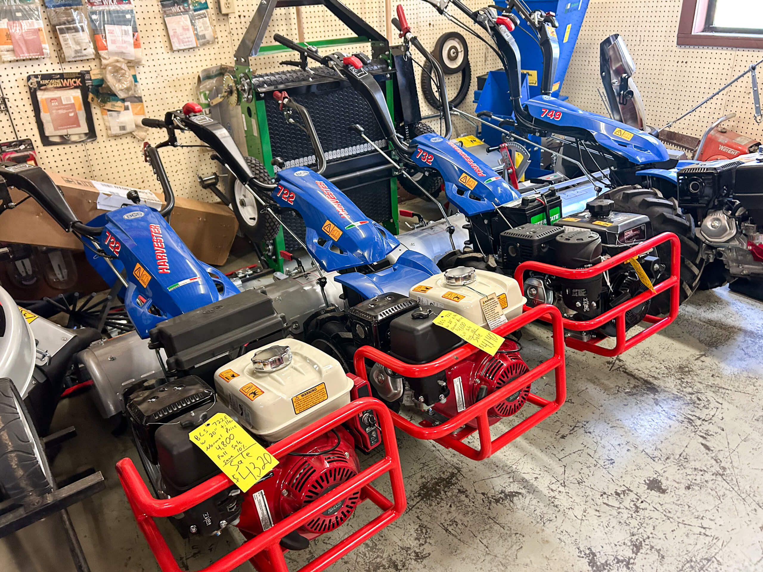 AK Small Engines Versailles, MO Several blue and red walk-behind tractors with yellow price tags are displayed in a store on a concrete floor, with equipment and tools hanging on the wall behind them.