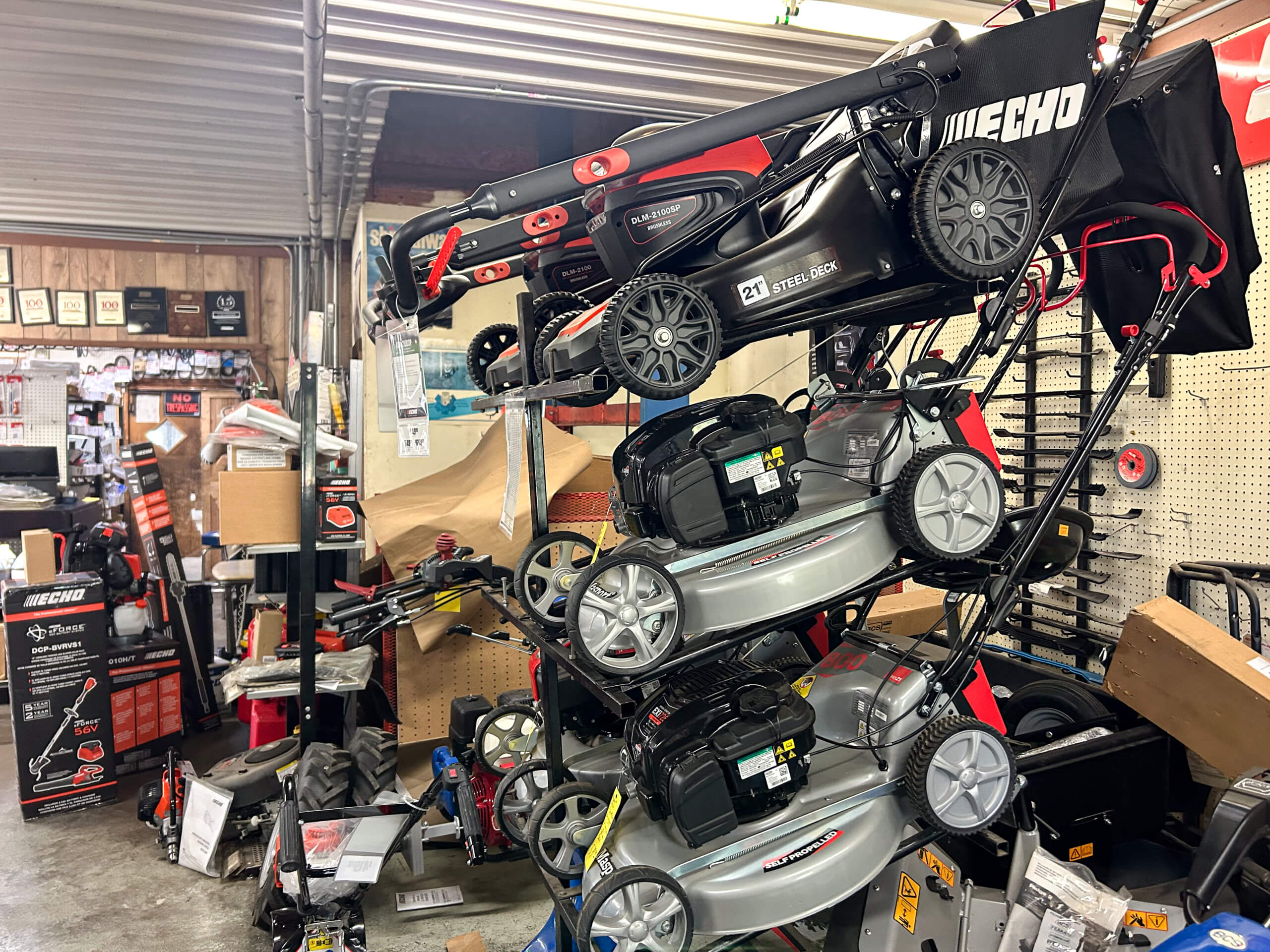 AK Small Engines Versailles, MO Several lawnmowers are stacked vertically on display inside a cluttered hardware or equipment store, with various tools and products visible in the background.