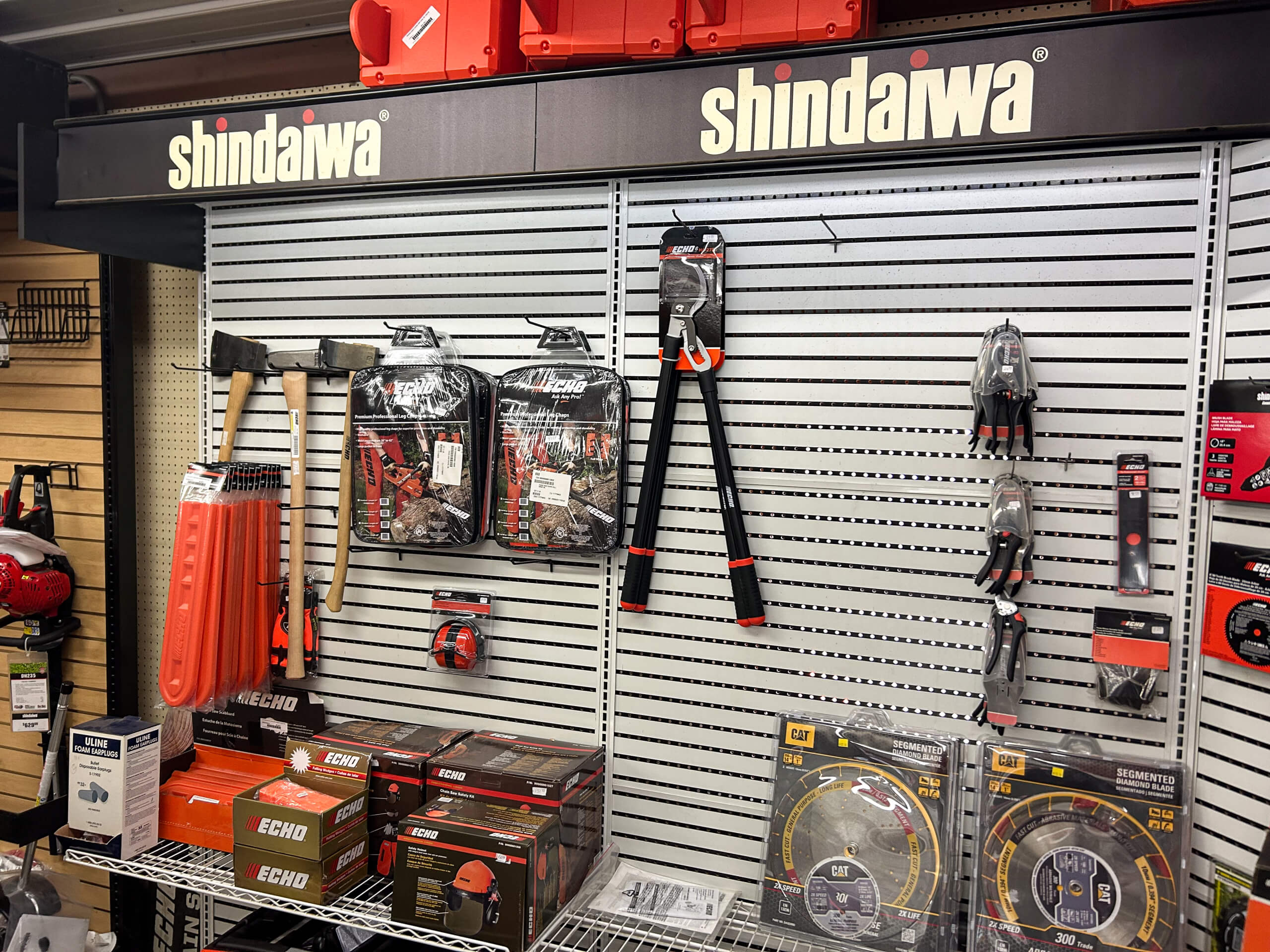 AK Small Engines Versailles, MO Display of Shindaiwa branded tools and accessories, including axes, loppers, gloves, and packaged equipment, arranged on a slatwall in a hardware store.