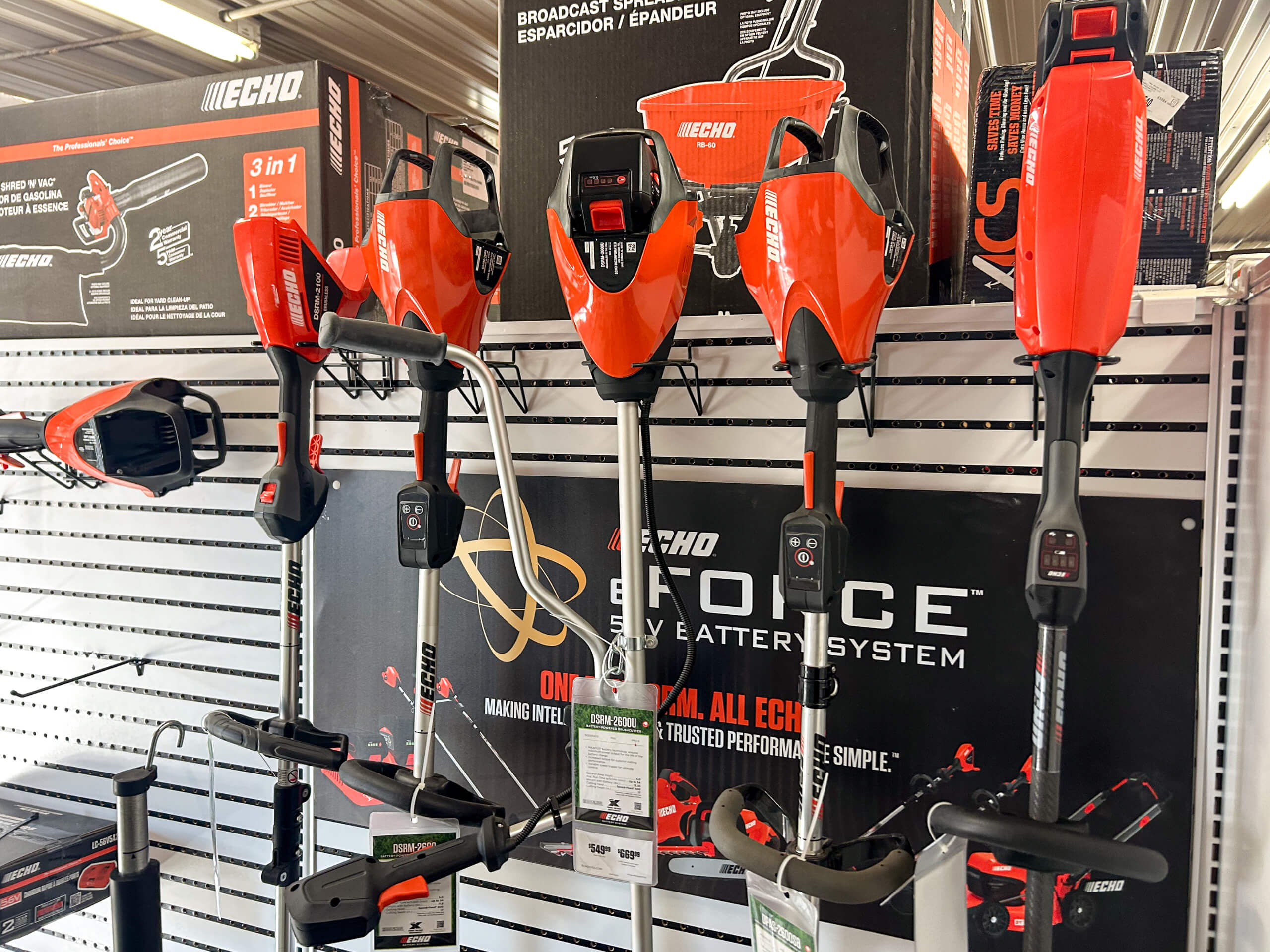 AK Small Engines Versailles, MO A row of red and black battery-powered string trimmers and blowers are displayed on a store rack with product boxes and signage above and behind them.