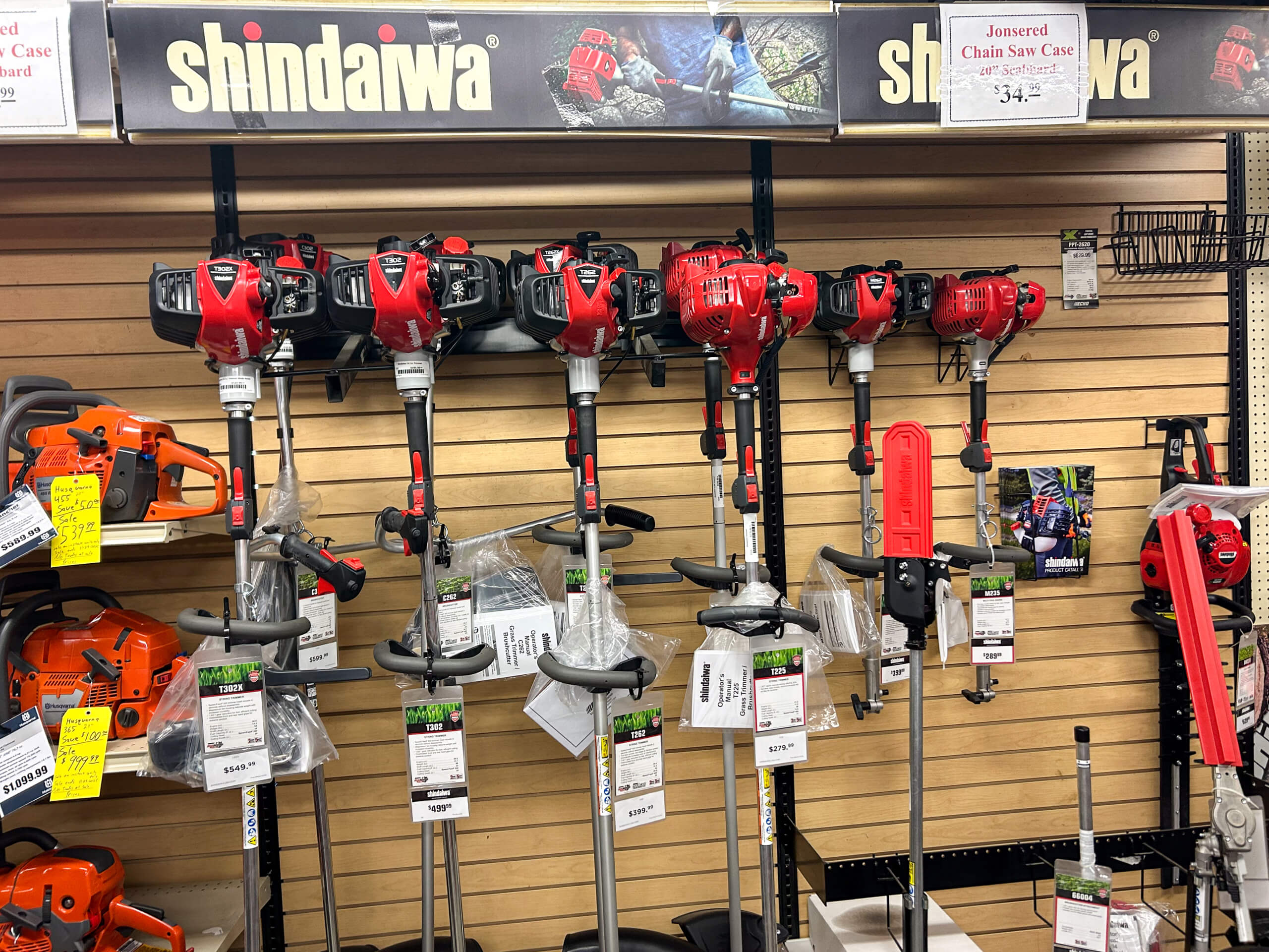 AK Small Engines Versailles, MO Multiple red and black string trimmers are displayed on a store wall rack, with orange chainsaws and accessories arranged below on the shelves.