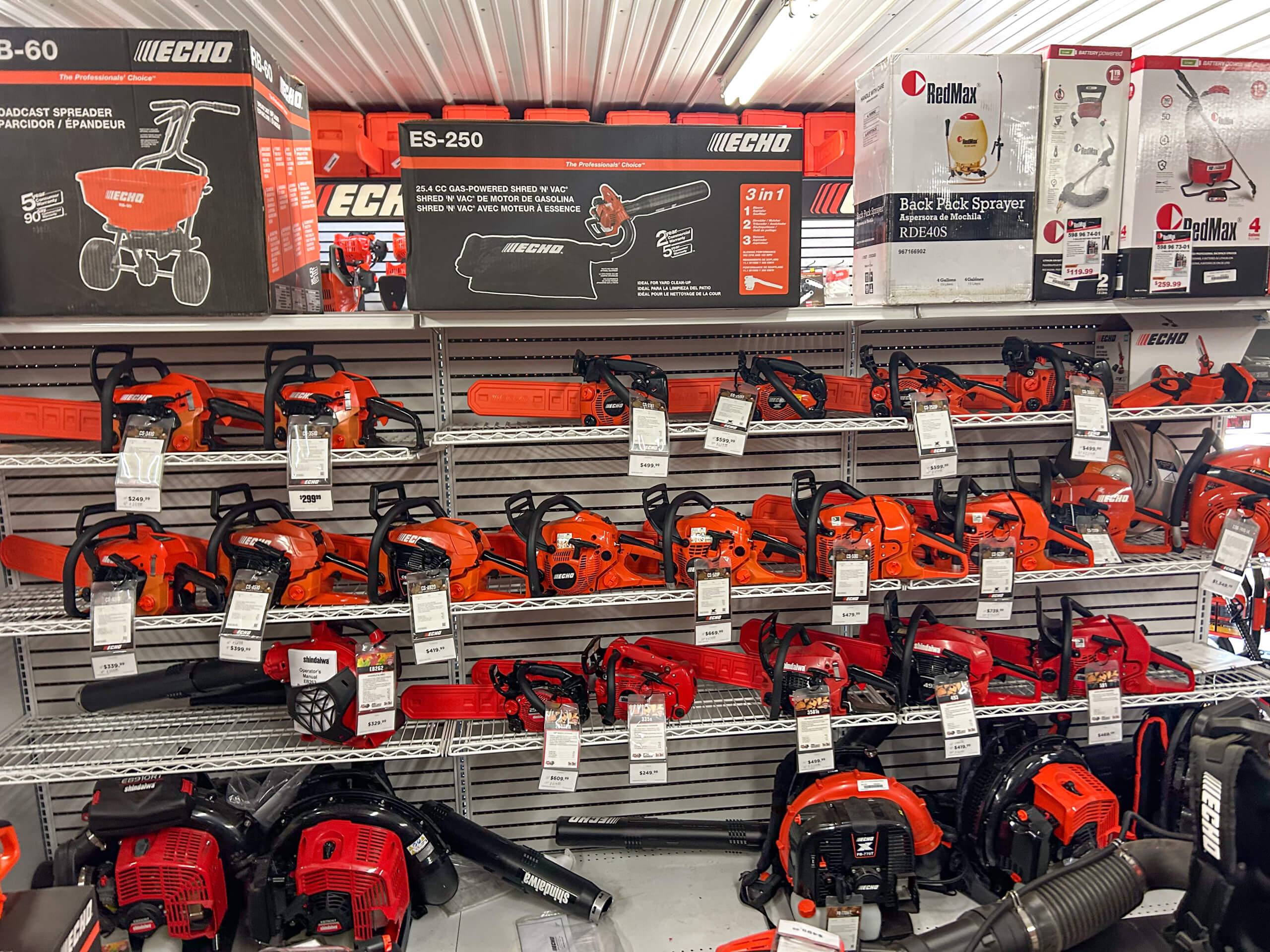 AK Small Engines Versailles, MO Shelves display various models of orange and red chainsaws, blowers, and yard equipment with price tags in a hardware store aisle.
