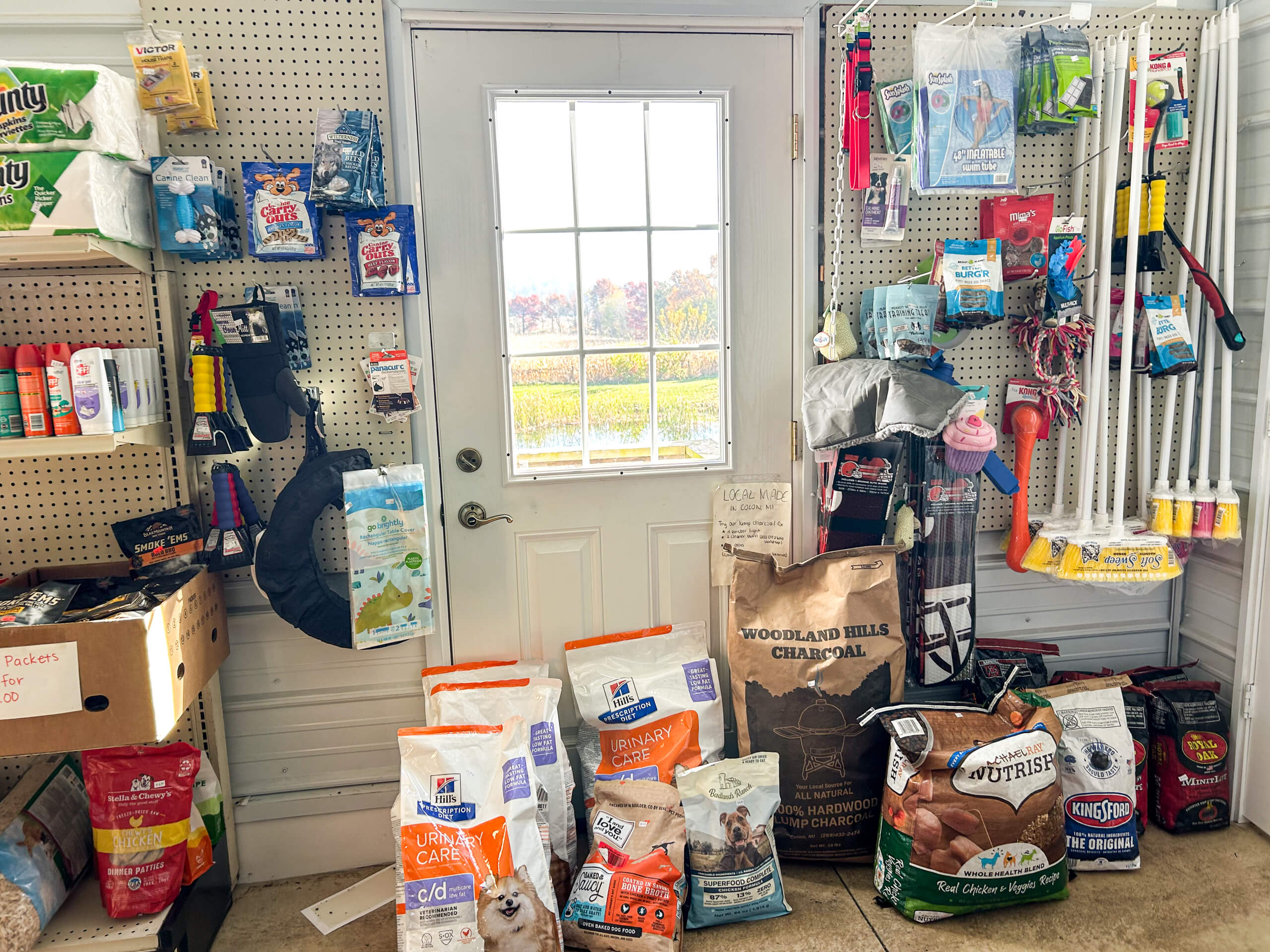 Sunrise Discount Grocery Store Colon MI Pet supplies, including food bags and accessories, are displayed on shelves and the floor in front of a white door with a window, inside a brightly lit room.
