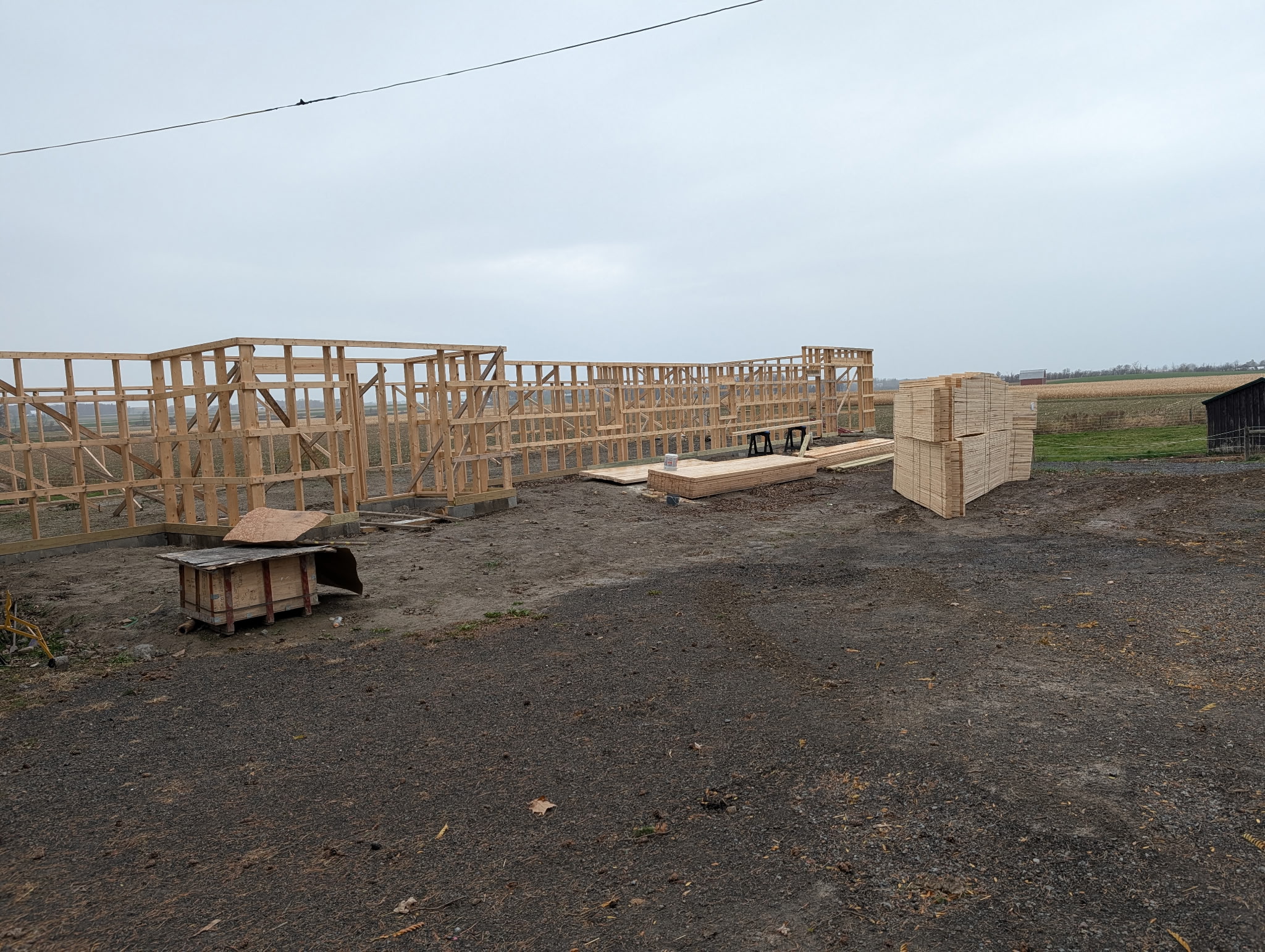 Wooden framework of a building under construction on a dirt lot, with stacks of lumber and open fields in the background.