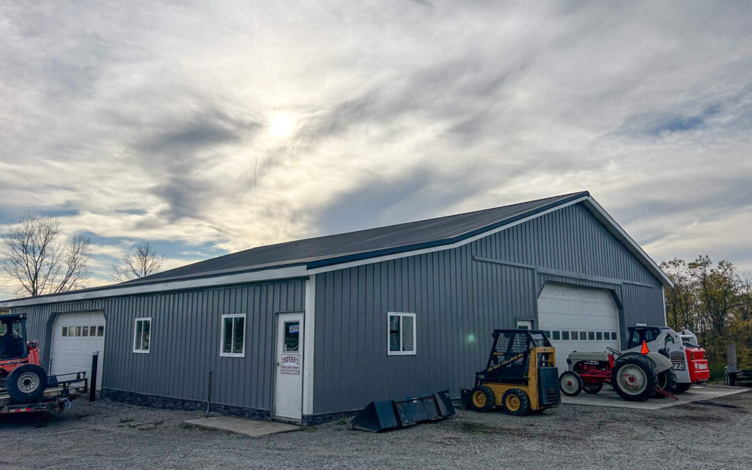 Troyer's Skid Loader Repair Rushsylvania, Ohio A gray metal building with large garage doors, surrounded by gravel and several pieces of construction equipment parked outside.