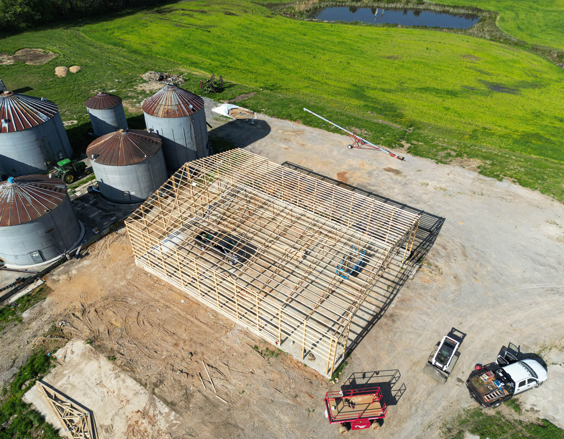 12.11.25 Mid-South Truss and Supply Gallery 1 Aerial view of a large barn under construction with wooden framing near several metal silos, a pond, vehicles, and green fields.