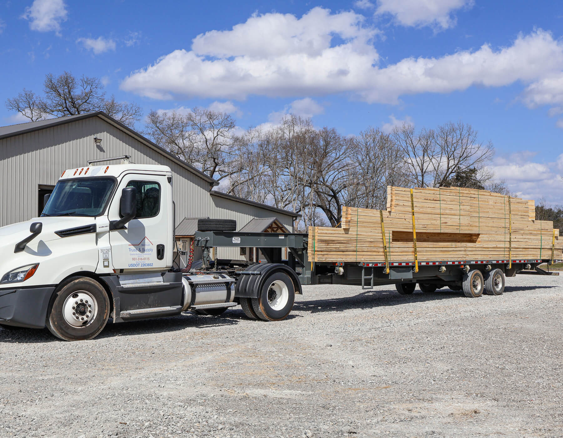 12.11.25 Mid-South Truss and Supply Gallery 3 A white semi-truck with a flatbed trailer loaded with stacked lumber is parked on a gravel lot near a metal building.
