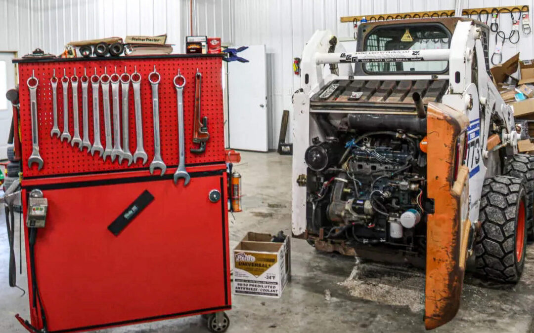 Troyer's Skidloader Repair Rushsylvania, OH A red tool chest with wrenches stands beside a skid steer loader with its rear engine compartment open inside a workshop.
