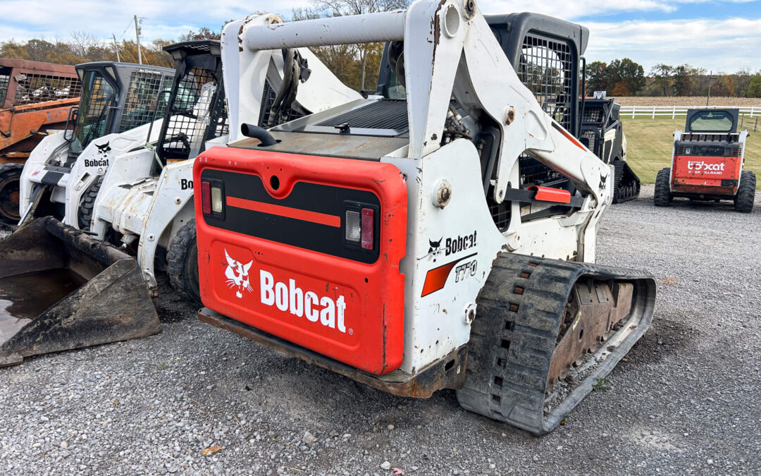 Troyer's Skidloader Repair Rushsylvania, OH A Bobcat compact track loader is parked on gravel beside other similar machines, with trees and a white fence in the background.