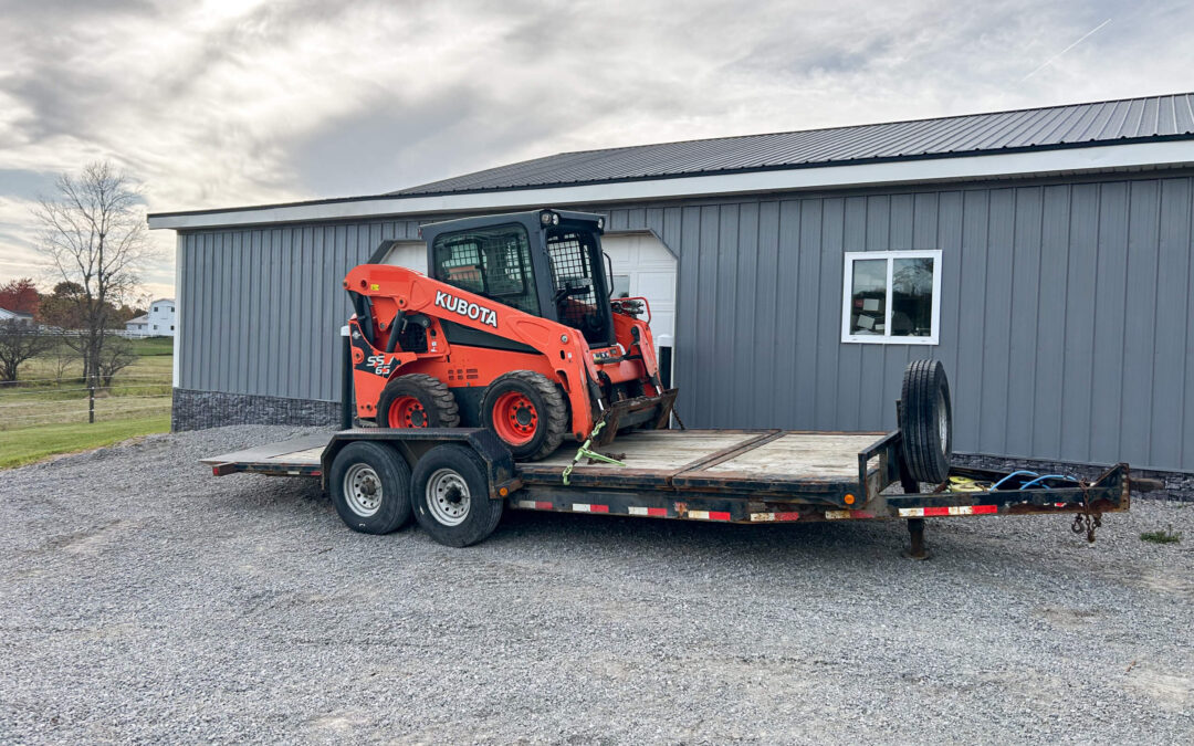 Troyer's Skidloader Repair Rushsylvania, OH An orange Kubota skid steer is parked on a flatbed trailer in front of a gray metal building on a gravel surface.