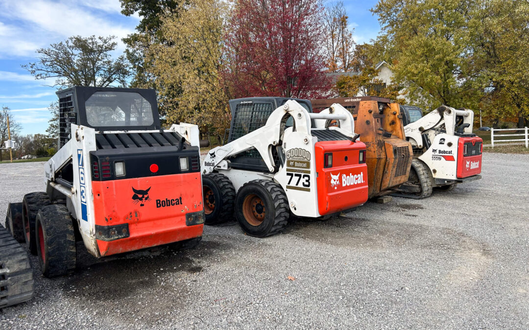 Troyer's Skidloader Repair Rushsylvania, OH Four Bobcat skid steer loaders are parked in a row on a gravel lot, with autumn trees in the background.