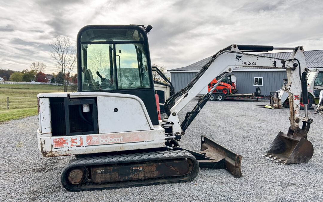Troyer's Skidloader Repair Rushsylvania, OH A white Bobcat mini excavator with tracks and bucket attachment is parked on gravel near a metal building and grassy field under a cloudy sky.