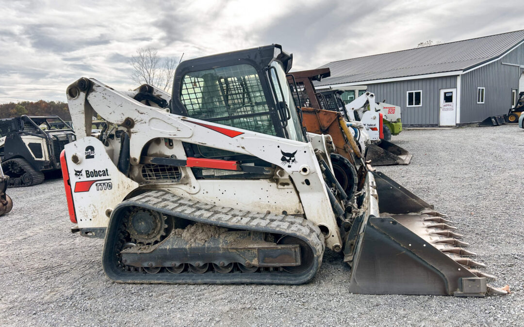 Troyer's Skidloader Repair Rushsylvania, OH A white Bobcat T770 compact track loader with a front bucket attachment is parked on a gravel lot near a gray industrial building.