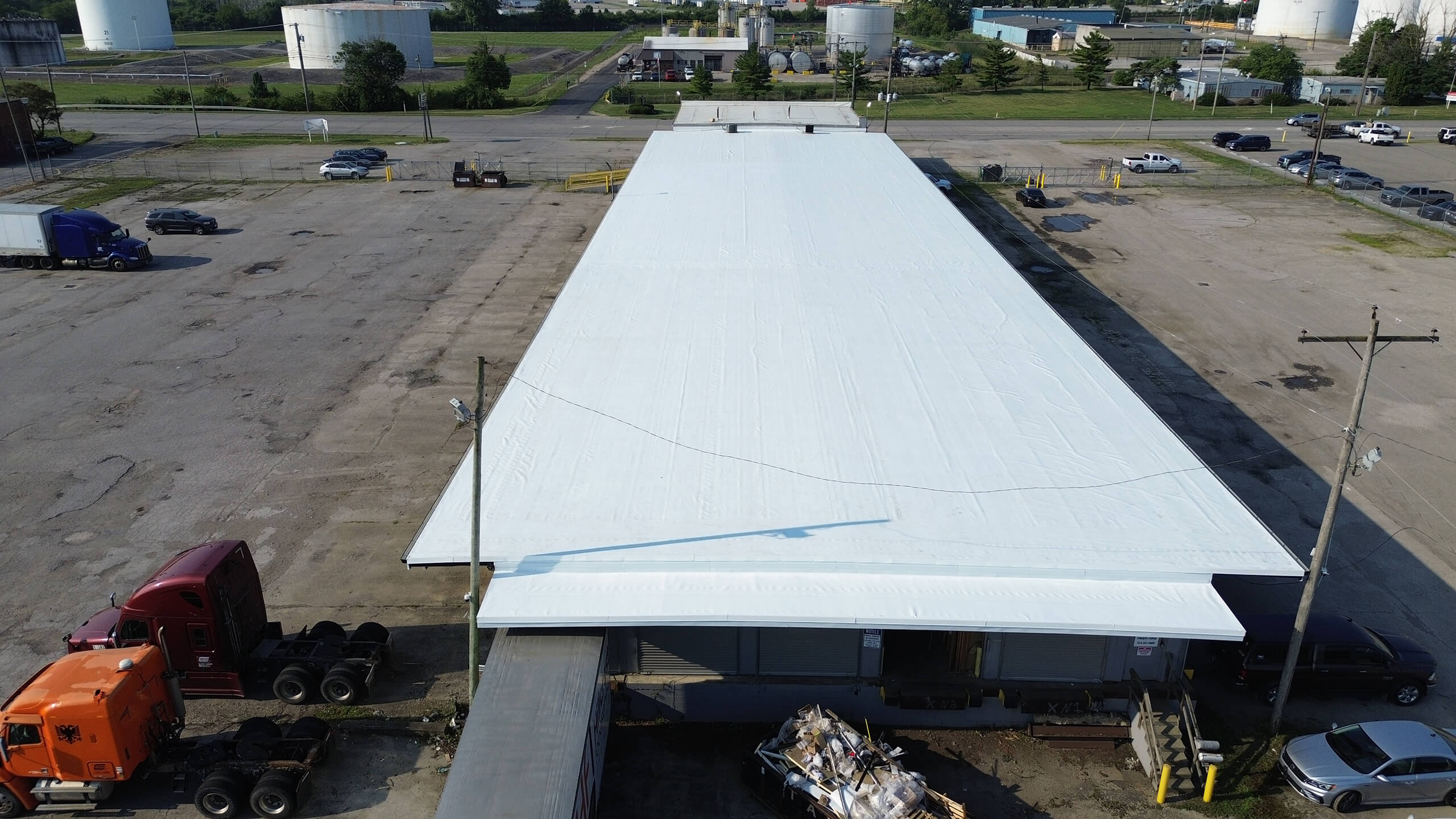 Aerial view of a large industrial warehouse with a white roof, surrounded by trucks, parked cars, and paved lot areas on a clear day.