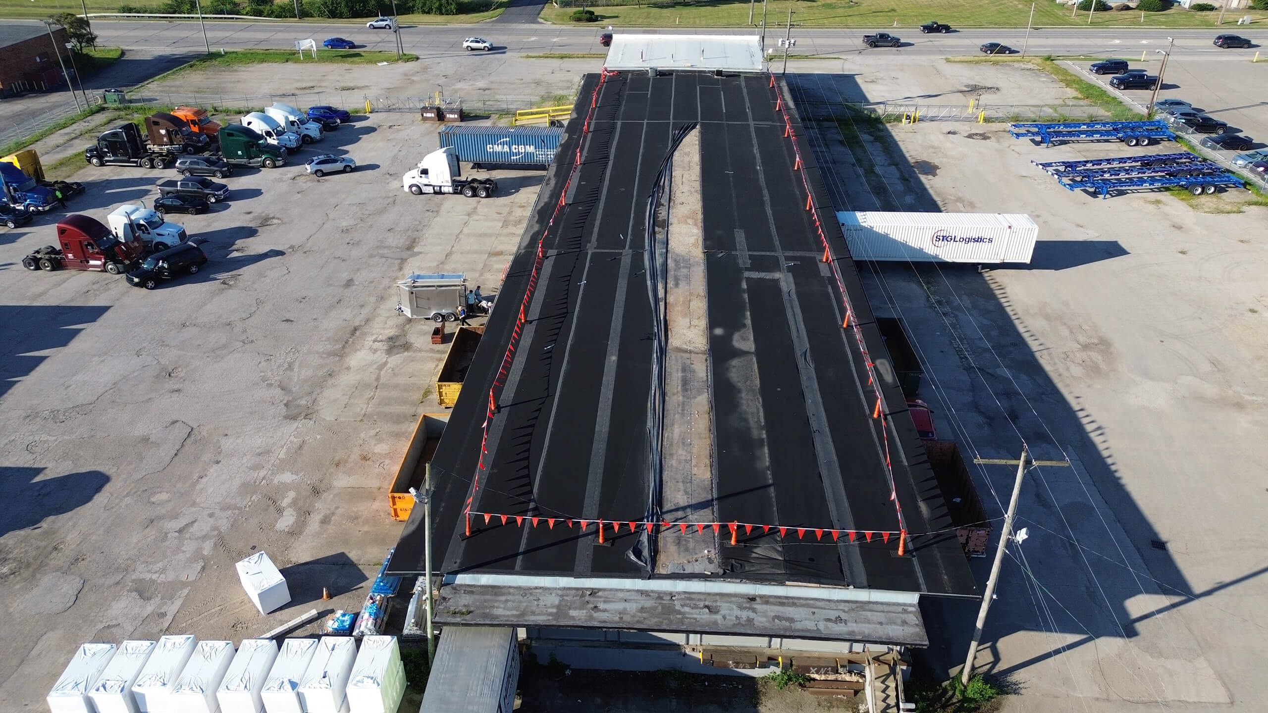 Aerial view of a large industrial building with a newly paved black roof, surrounded by trucks, containers, and parking areas.