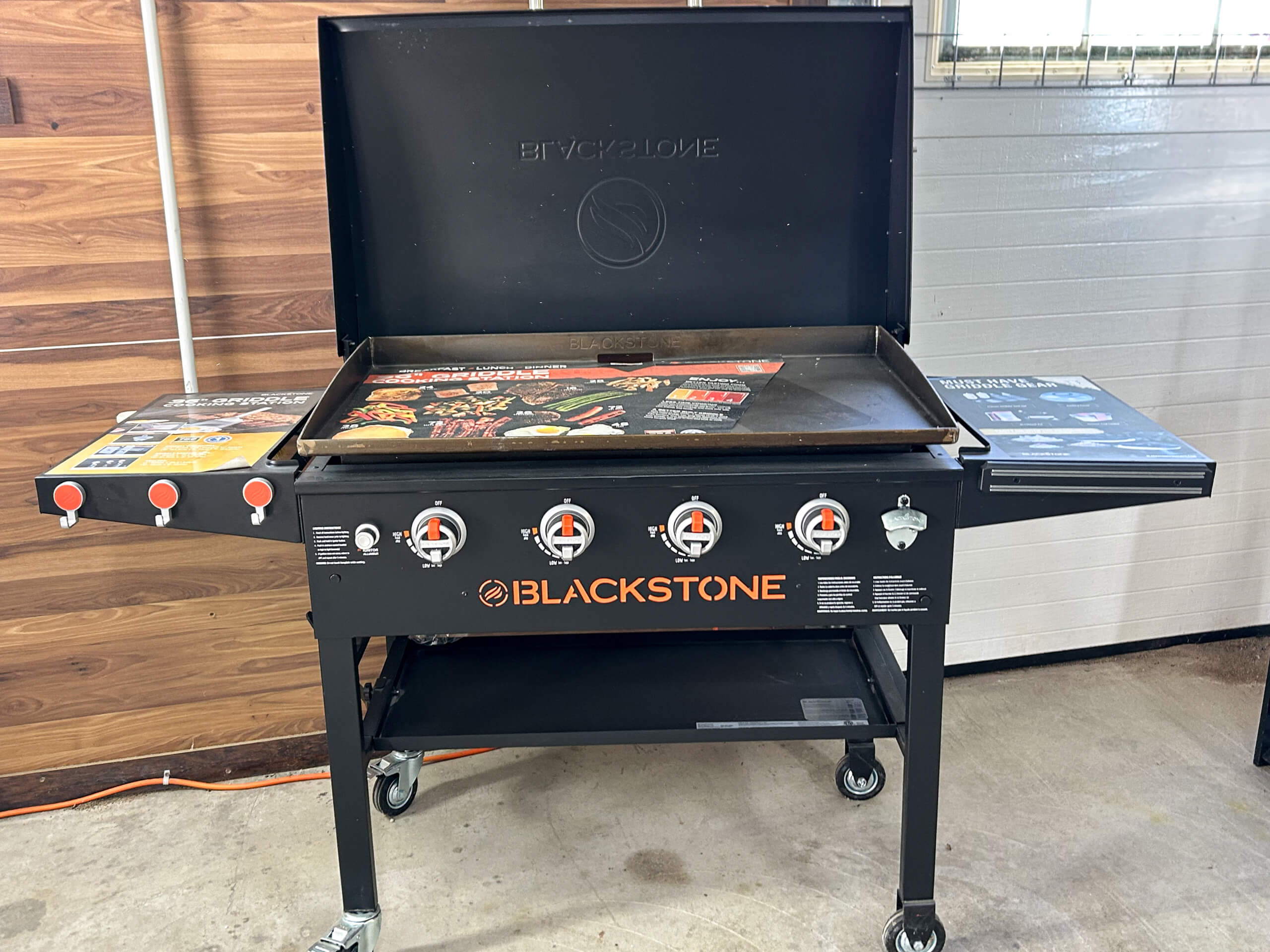 Graber Farm & Home Center Odon Indiana A Blackstone propane griddle with four burners, side shelves, and a closed lid, positioned indoors on a concrete floor.