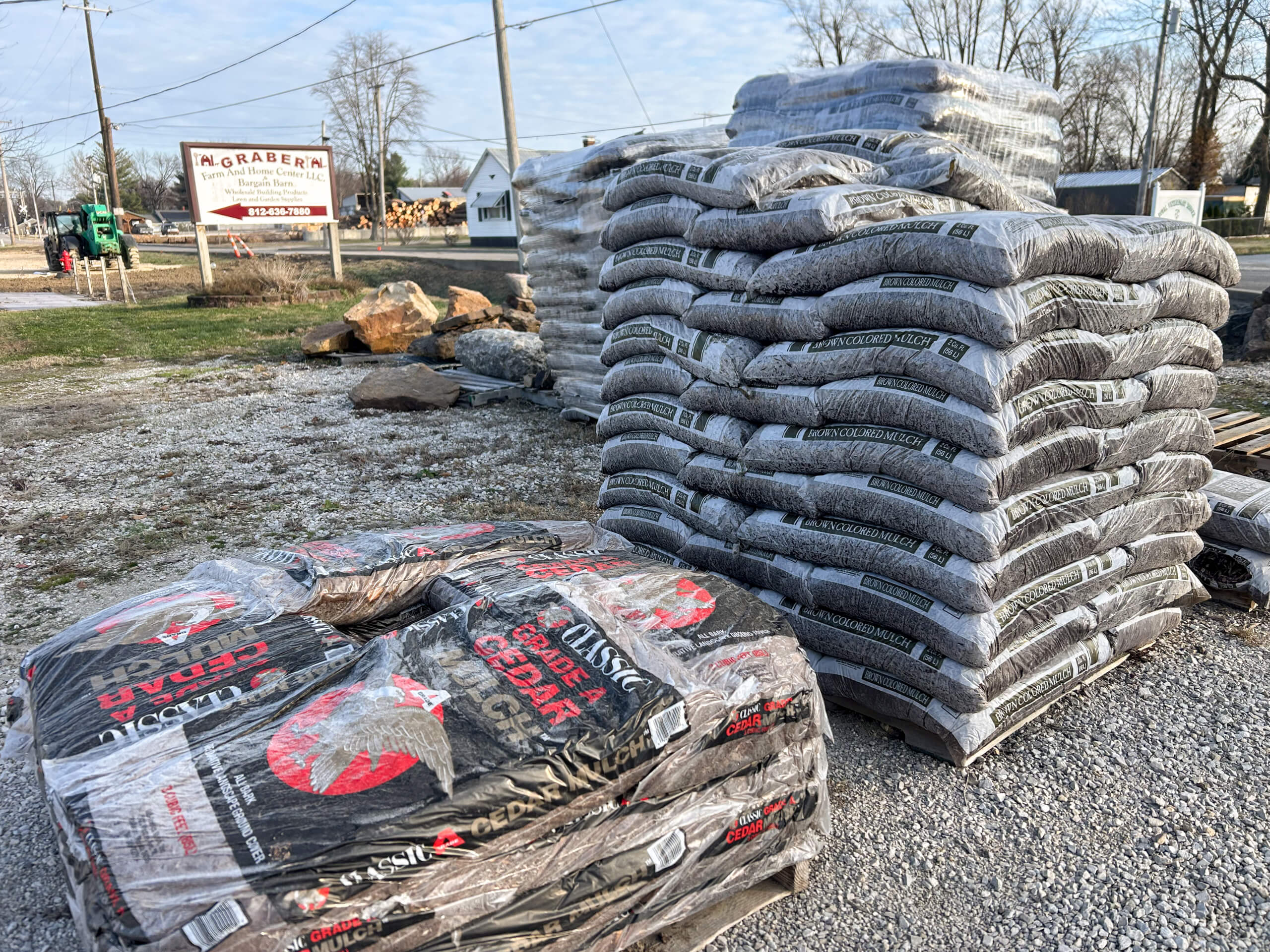 Graber Farm & Home Center Odon Indiana Several stacks of bagged mulch or soil sit on a gravel lot outside, near a sign reading "M. GRABERNA." Trees and a few buildings are visible in the background.