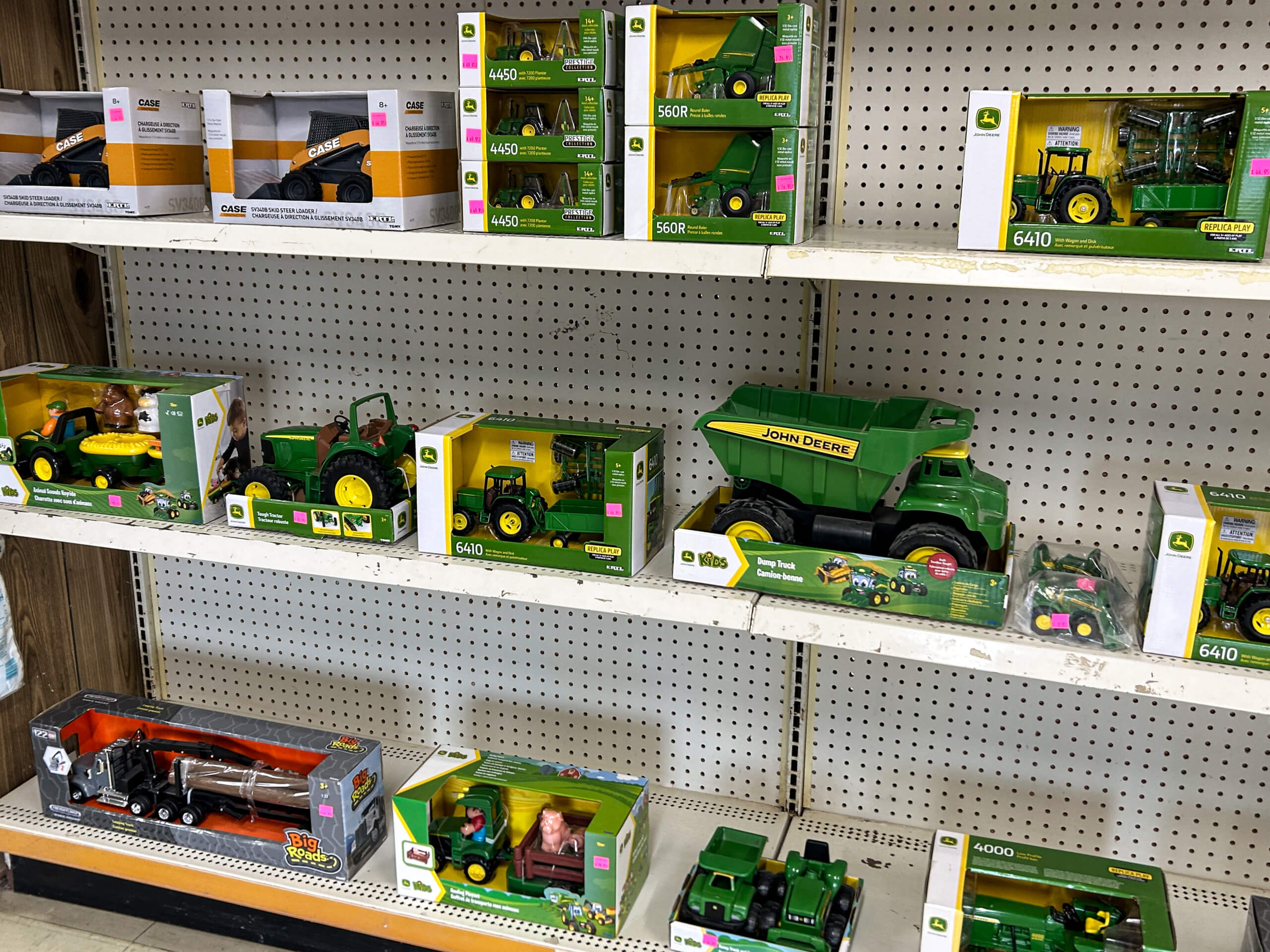 Graber Farm & Home Center Odon Indiana Shelves display various John Deere toy tractors and trucks in green and yellow packaging, arranged neatly on a retail store shelf with price stickers visible.