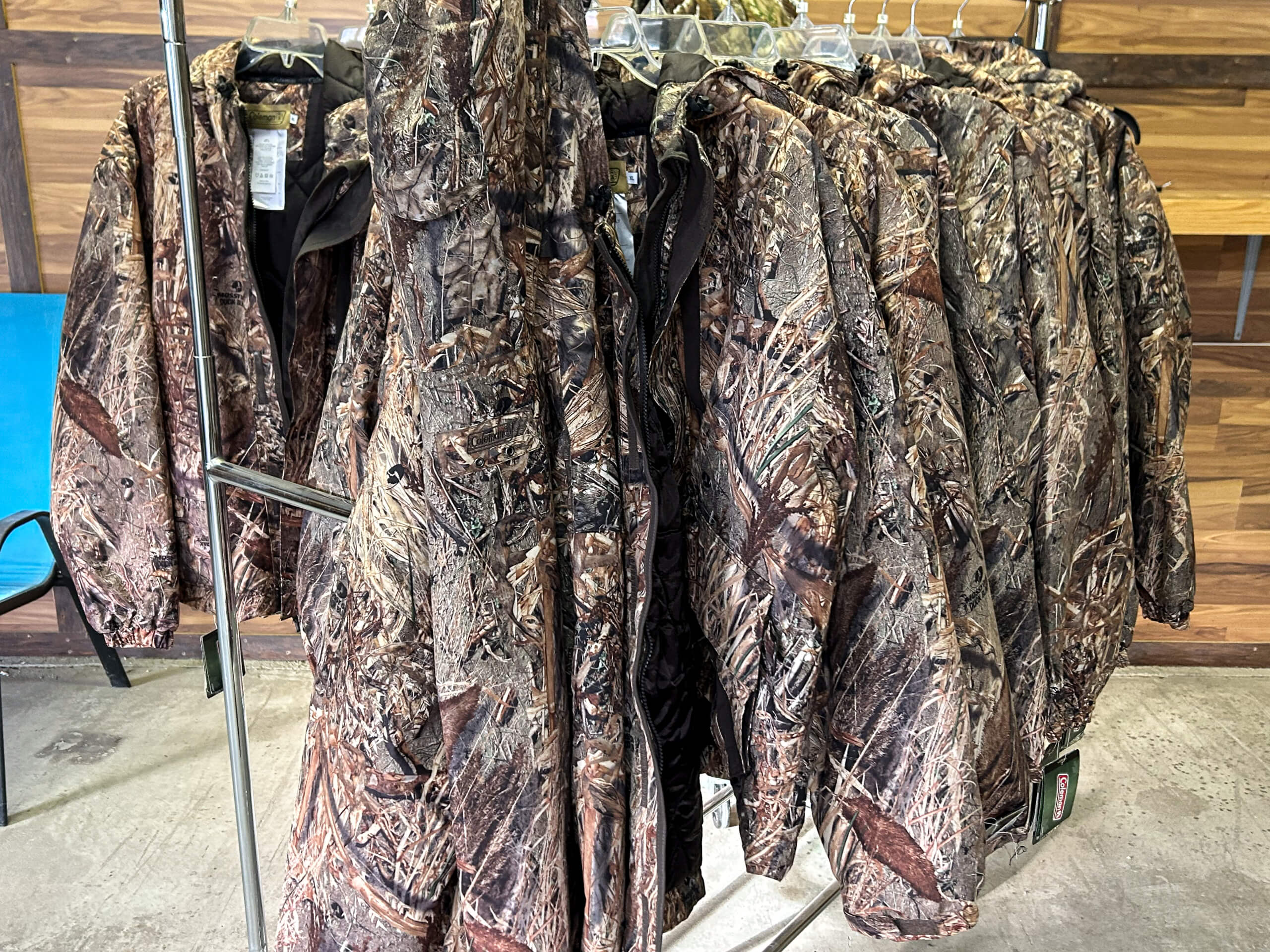 Graber Farm & Home Center Odon Indiana Several camouflage jackets hang on a metal rack indoors, with a wood-paneled wall and blue chair visible in the background.
