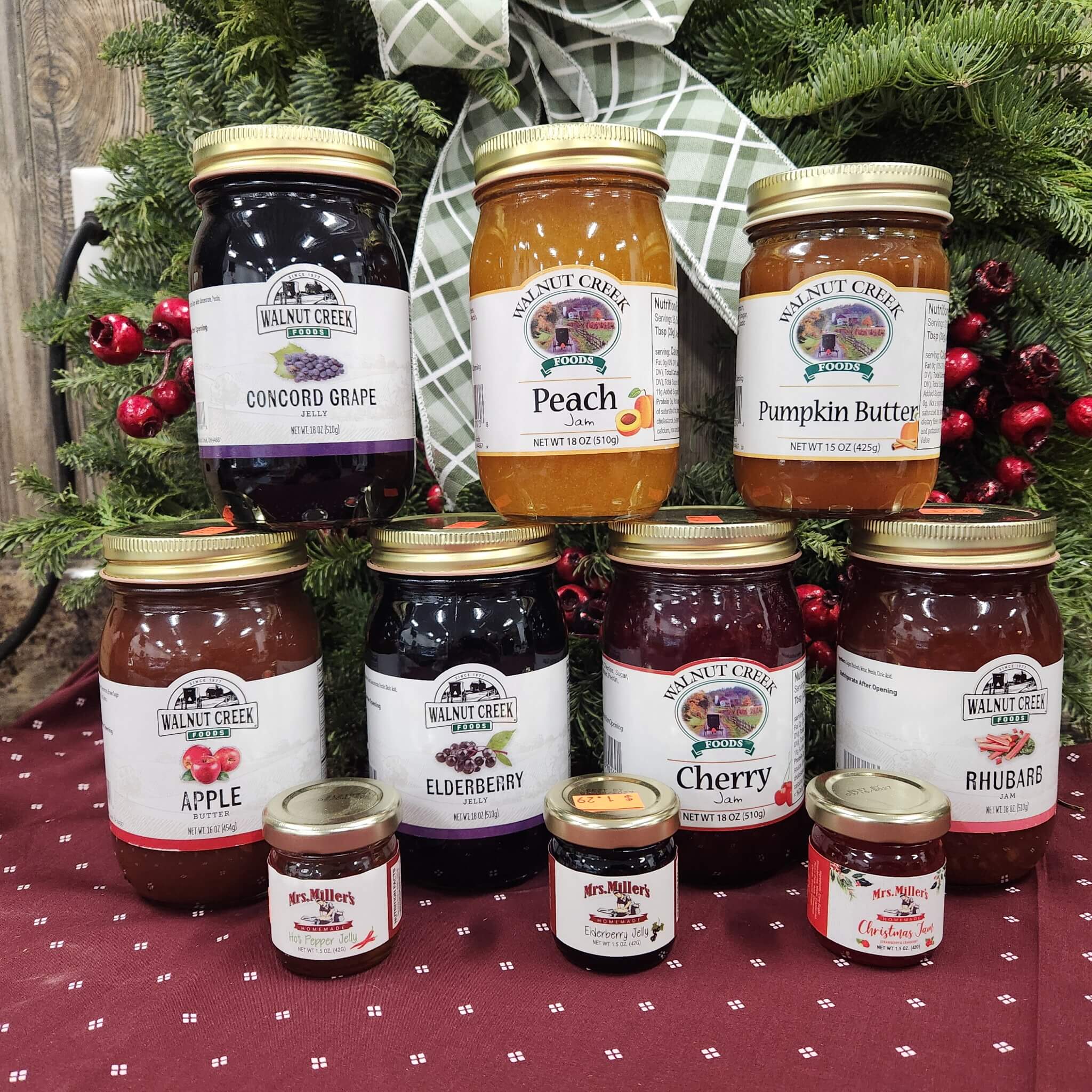 Countrty Pantry Sheridan, MI Nine jars of Walnut Creek preserves and three small jars of Mrs. Miller's jams are arranged on a red cloth in front of a decorated Christmas wreath.