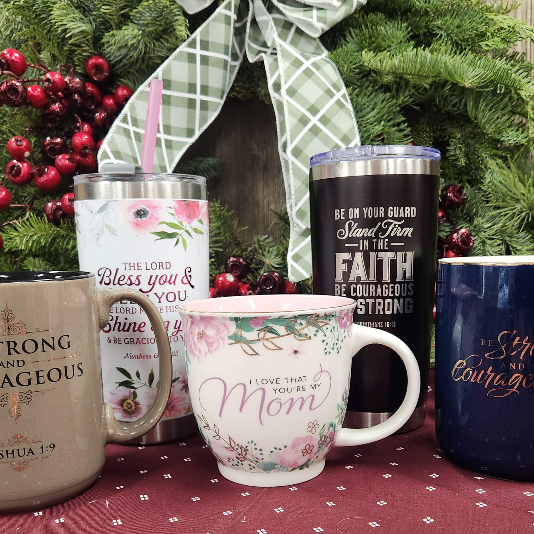 Countrty Pantry Sheridan, MI Five mugs and tumblers with inspirational quotes are arranged on a red tablecloth in front of a holiday wreath decorated with greenery, berries, and a plaid bow.