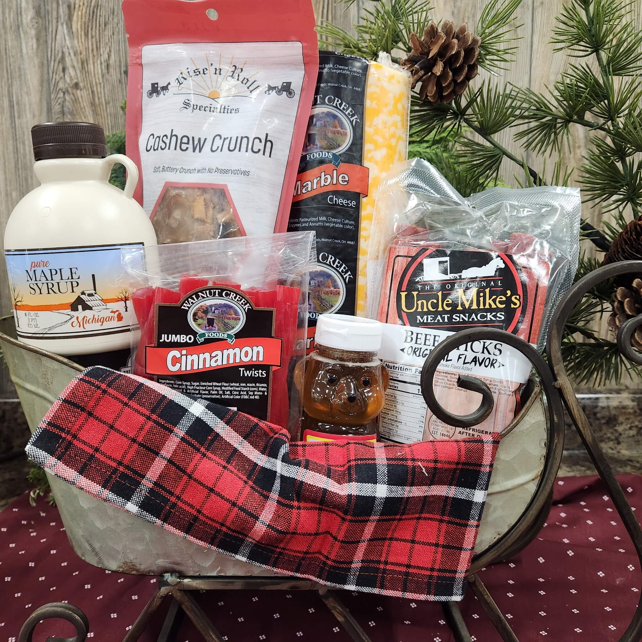Countrty Pantry Sheridan, MI A metal sleigh holds assorted snacks, including maple syrup, cashew crunch, cheese, cinnamon twists, honey, meat snacks, and a red plaid cloth, with pinecones and greenery in the background.
