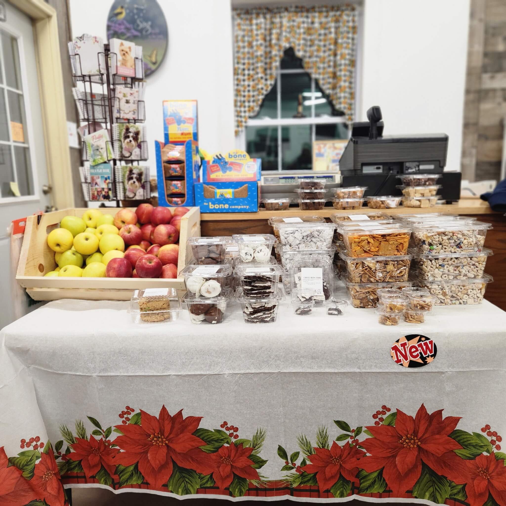 Countrty Pantry Sheridan, MI Table display with apples, assorted packaged snacks, and baked goods in front of a store counter decorated with a poinsettia tablecloth.