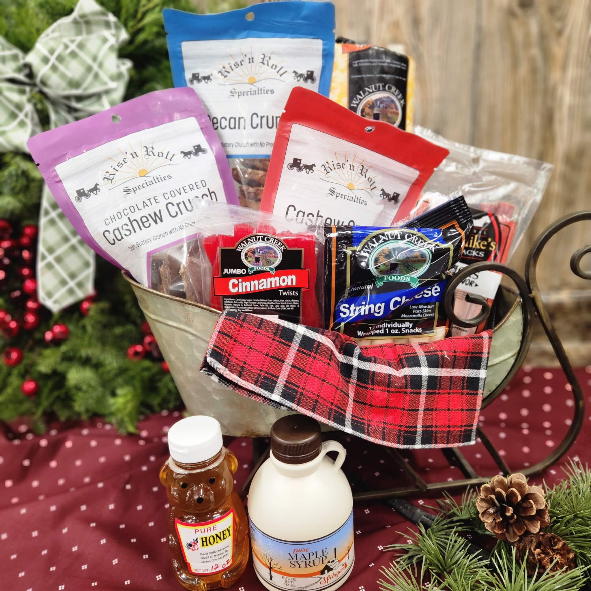 Countrty Pantry Sheridan, MI A gift basket contains assorted snack bags, honey, maple syrup, and cheese, displayed on a red cloth with festive greenery and a plaid napkin.