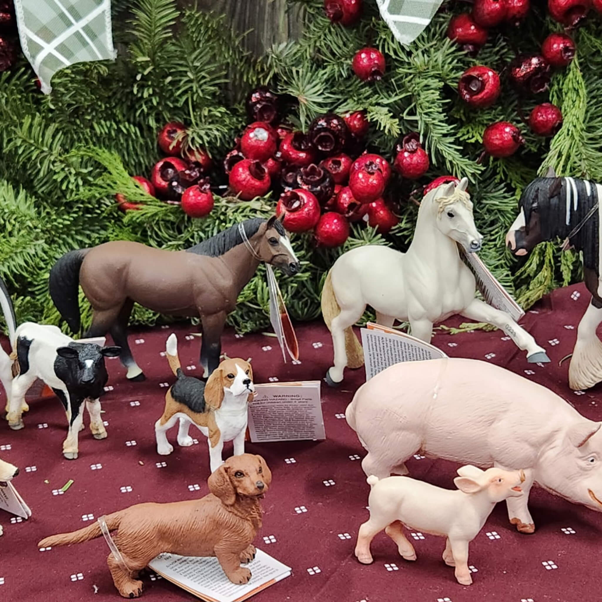 Countrty Pantry Sheridan, MI Assorted animal figurines, including a horse, cow, dog, pig, and goat, are displayed on a maroon cloth with green foliage and red berries in the background.