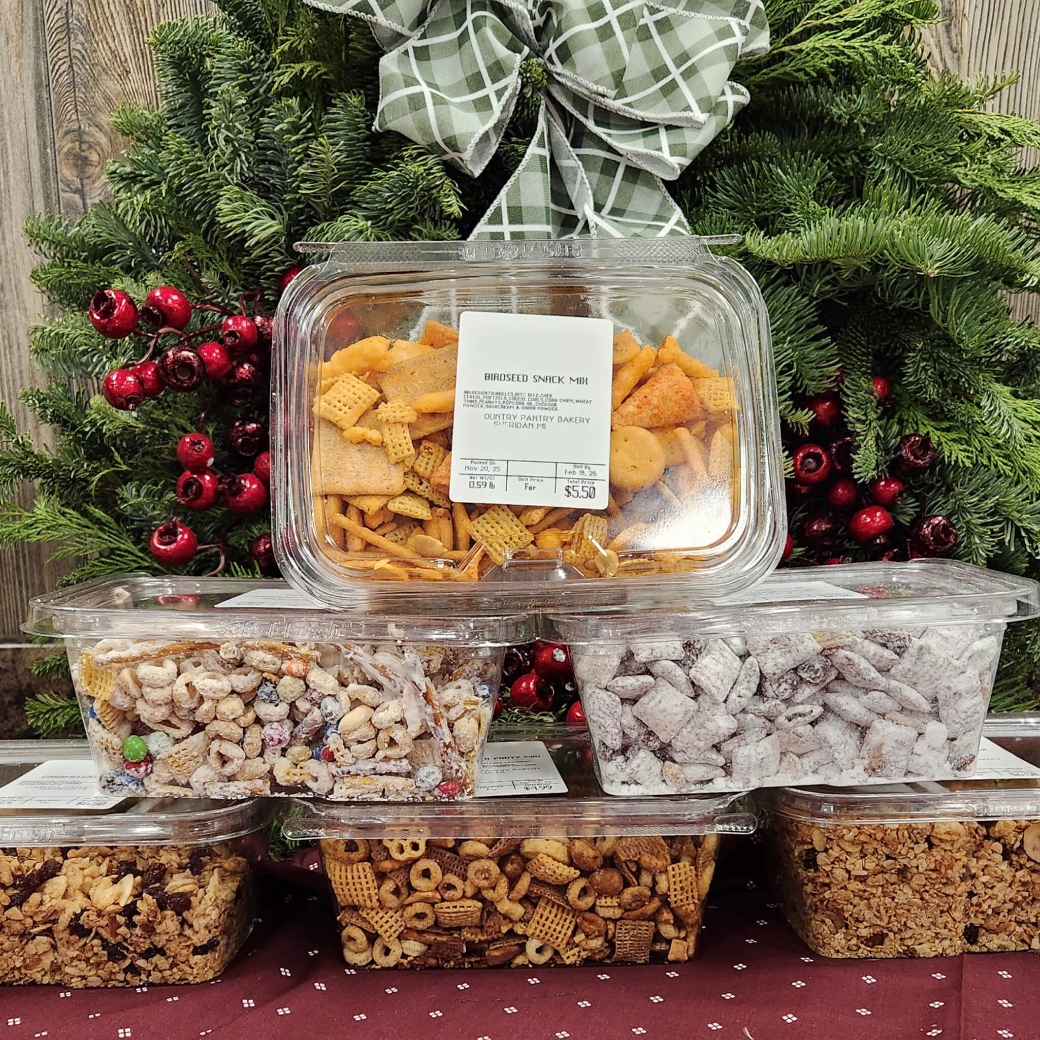 Countrty Pantry Sheridan, MI Assorted snack mixes in plastic containers are stacked in front of holiday greenery and ribbon, displayed on a red cloth with a snowflake pattern.