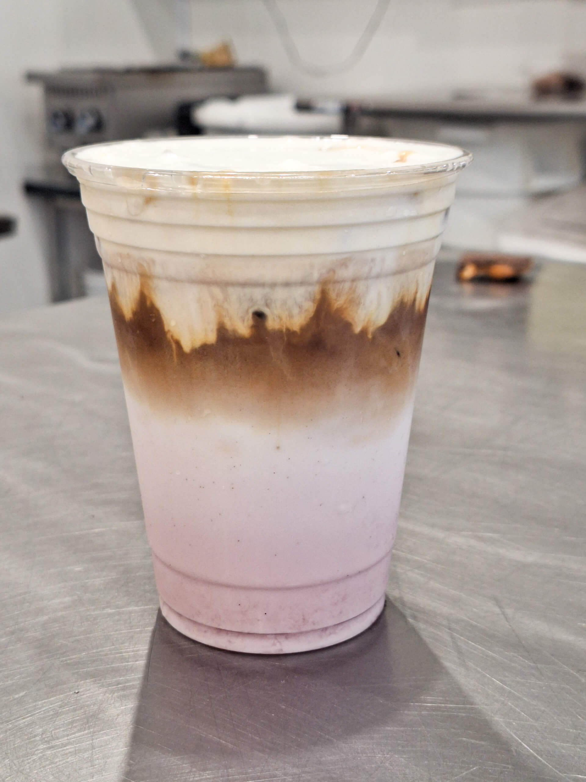 Dutch Heritage Bakery Sturgis MI A plastic cup filled with a layered iced drink, showing distinct pink, white, and brown layers, sits on a stainless steel countertop in a kitchen setting.