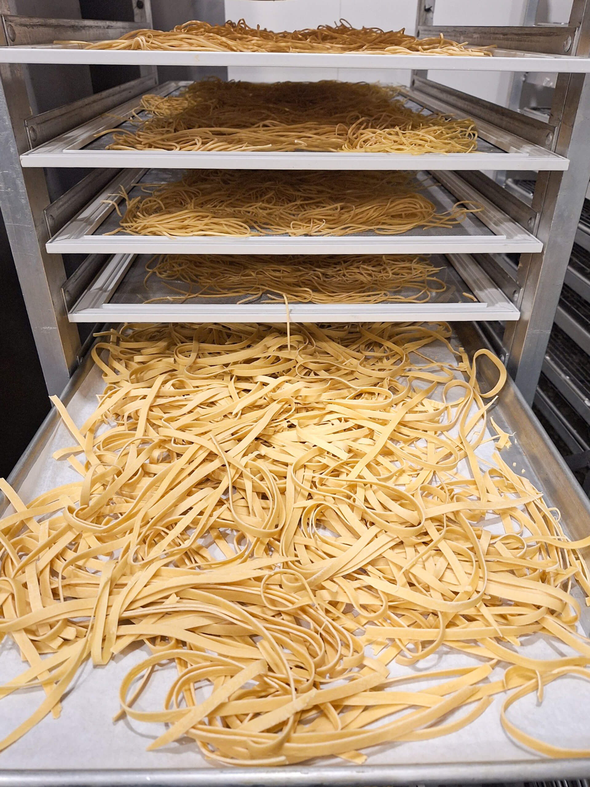 Dutch Heritage Bakery Sturgis MI Four trays of uncooked fettuccine pasta are stacked on a metal rack, with pasta spread out evenly on each tray.