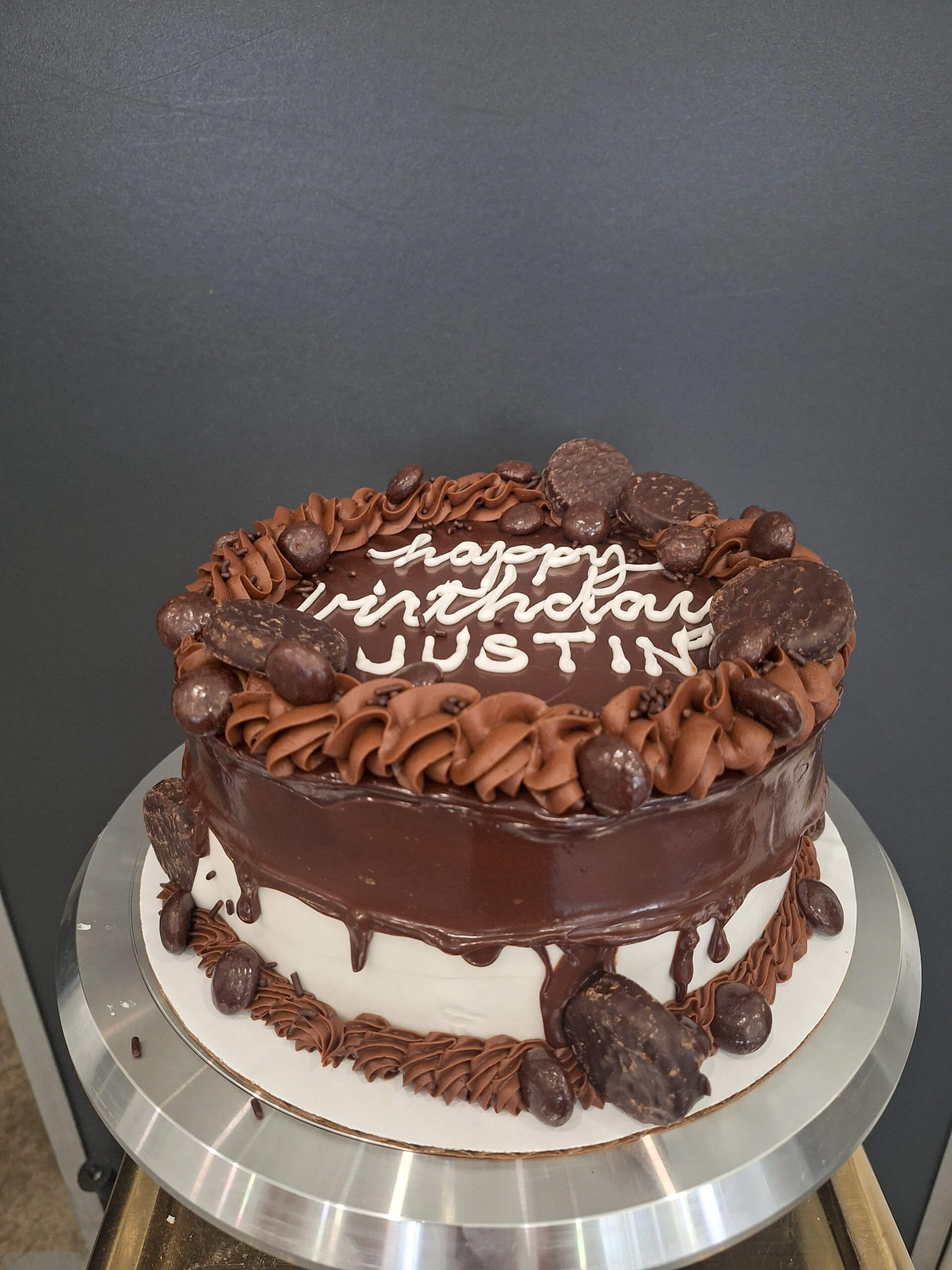 Dutch Heritage Bakery Sturgis MI A round chocolate and vanilla cake with chocolate frosting and decorations, featuring the text "happy birthday JUSTINE" written in white icing on top.