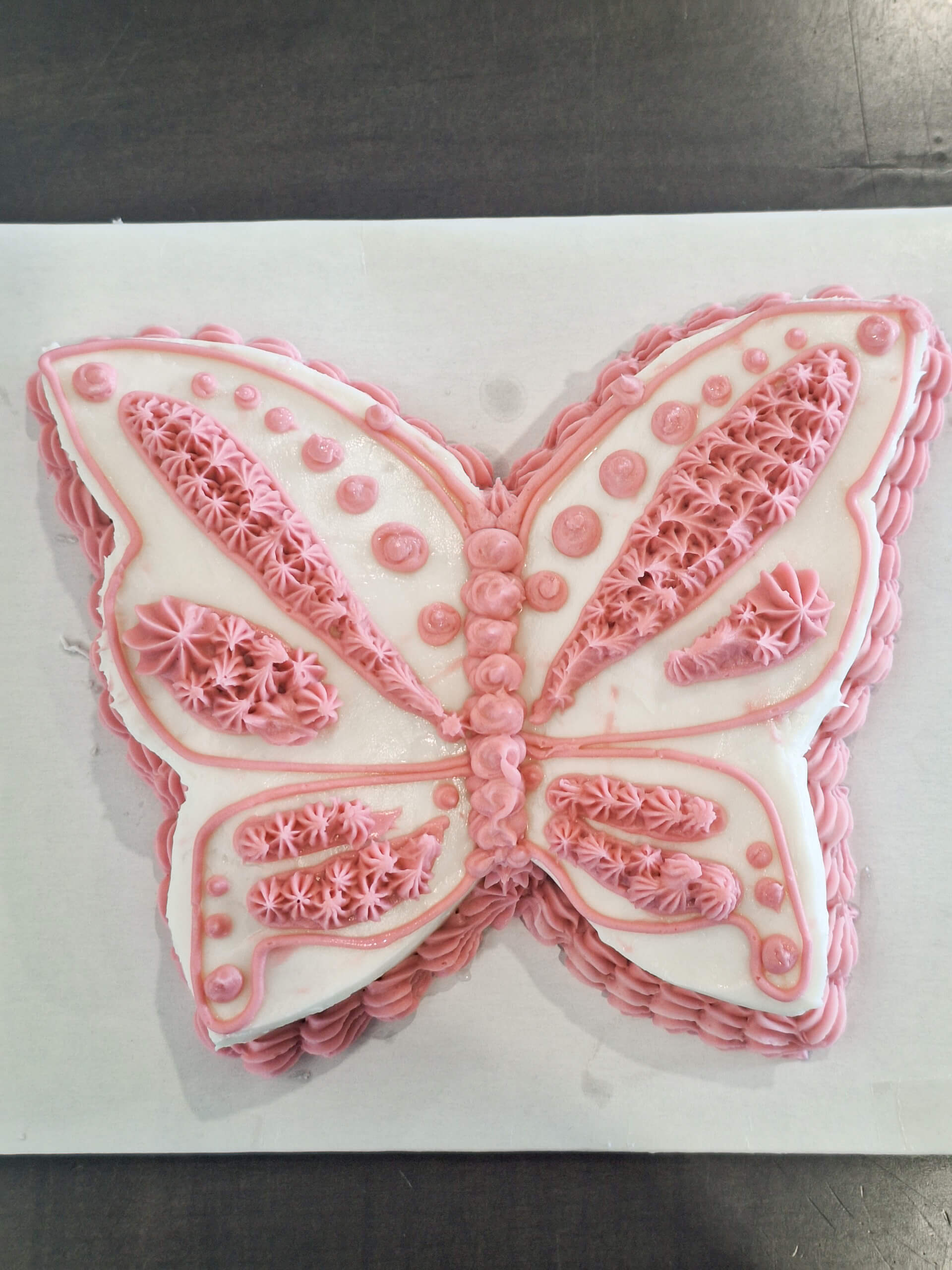 Dutch Heritage Bakery Sturgis MI A butterfly-shaped cake decorated with white icing and pink piped frosting patterns, displayed on a white rectangular board.