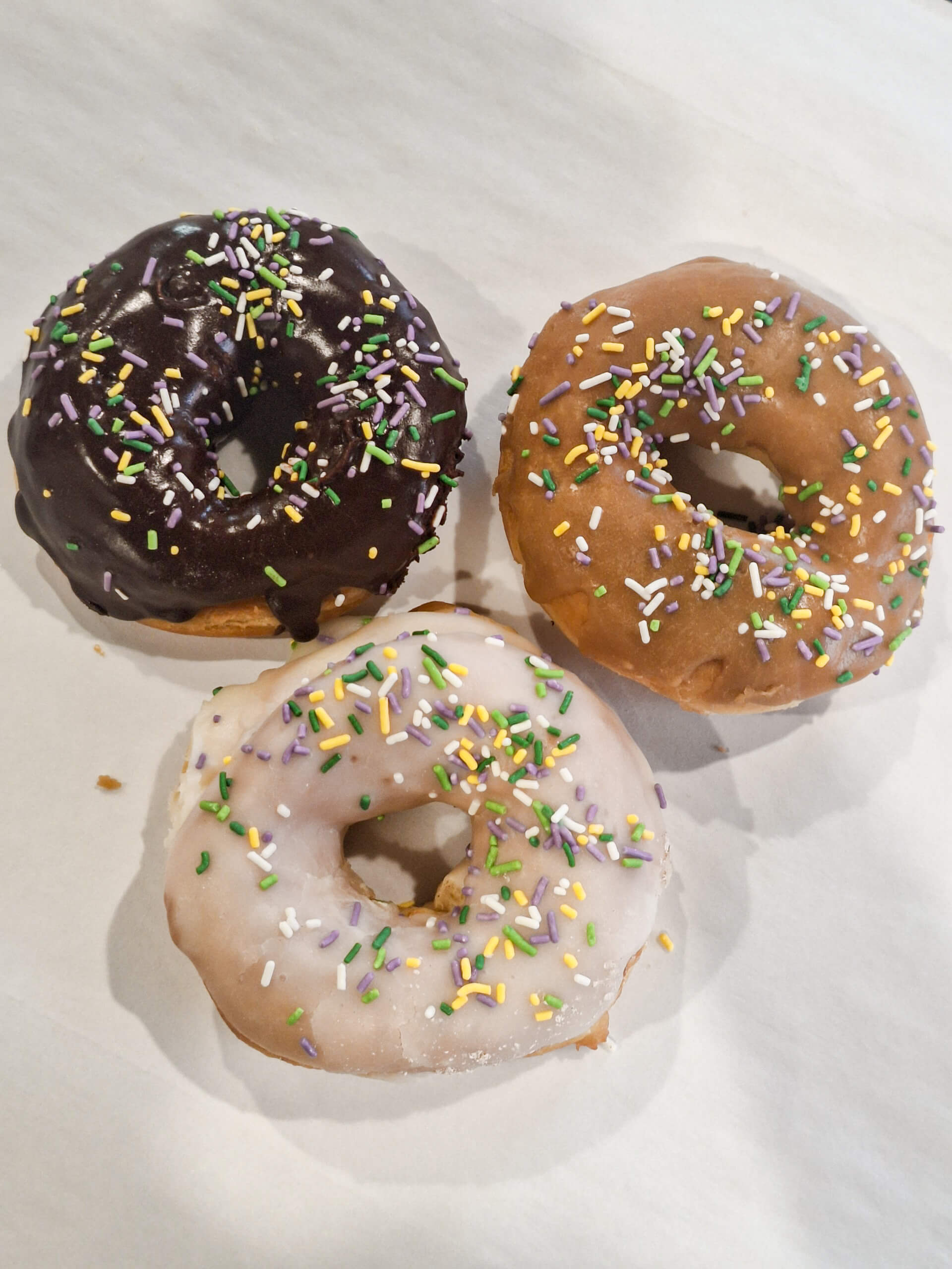 Dutch Heritage Bakery Sturgis MI Three donuts with chocolate, vanilla, and white icing, each topped with multicolored sprinkles, arranged on a white surface.
