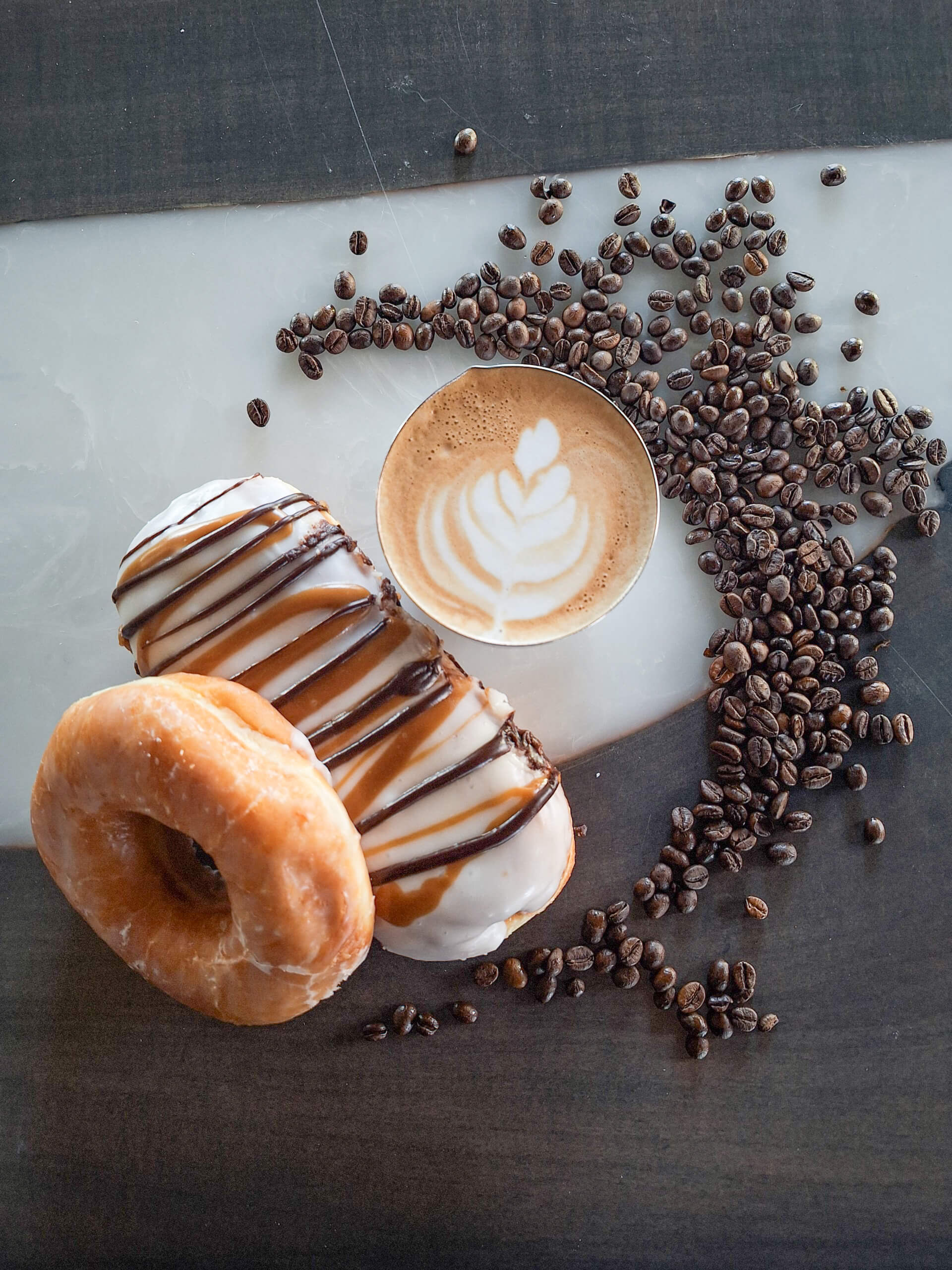 Dutch Heritage Bakery Sturgis MI A cup of latte with leaf latte art, two donuts—one glazed and one with white icing and chocolate drizzle—surrounded by scattered coffee beans on a dark surface.