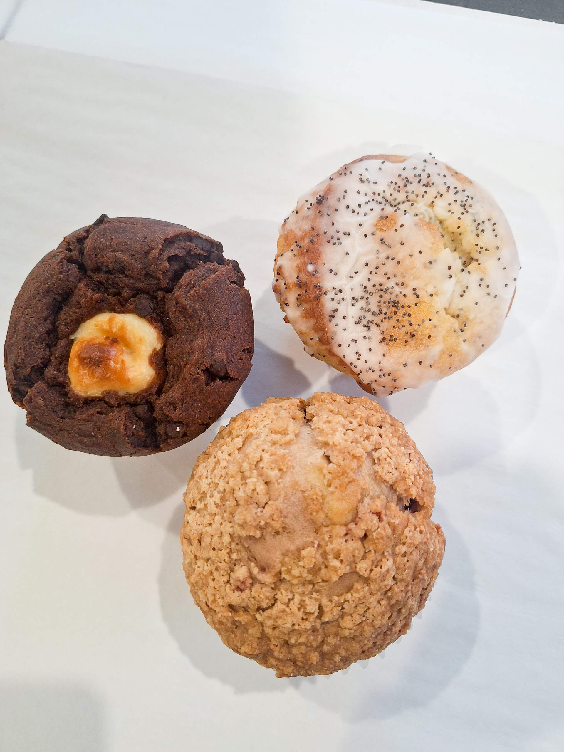 Dutch Heritage Bakery Sturgis MI Three muffins on a white surface: one chocolate with a marshmallow center, one topped with white glaze and poppy seeds, and one with a crumbly oat topping.