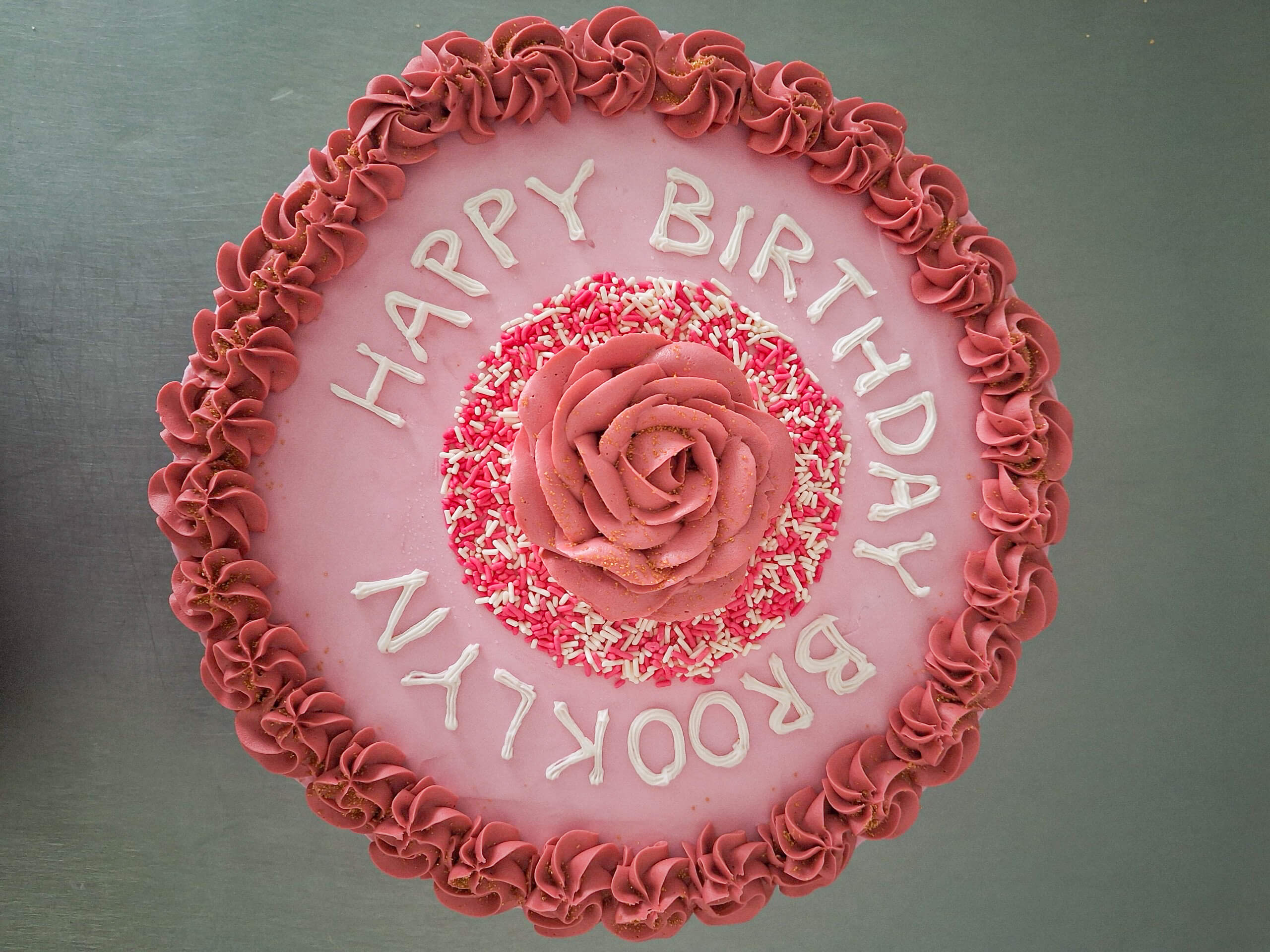 Dutch Heritage Bakery Sturgis MI A round pink cake with piped borders, a central rose decoration, and the words "HAPPY BIRTHDAY BROOKLYN" written in white icing.