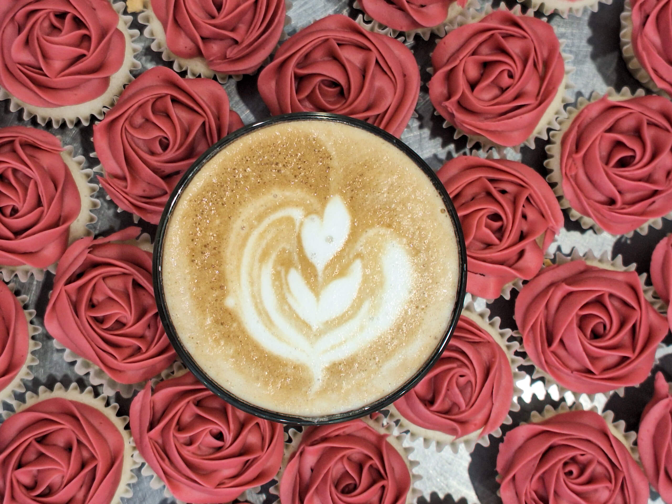 Dutch Heritage Bakery Sturgis MI A cup of latte with leaf latte art sits in the center, surrounded by cupcakes topped with pink rose-shaped frosting.